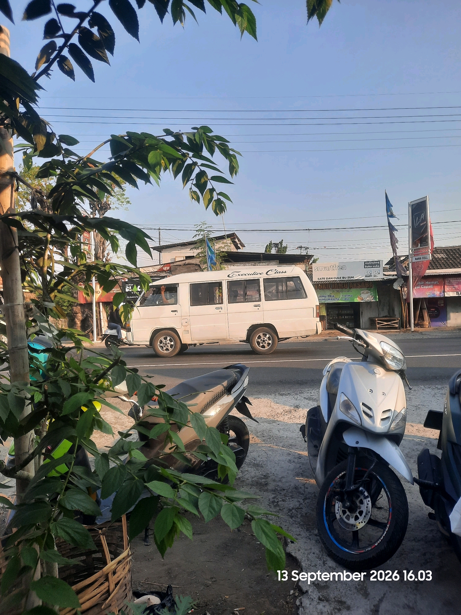 In Sukokerto, Indonesia, on September 13, 2026, at 4:03 PM, a white passenger van labeled "Executive Class" drives down a street lined with buildings and overhead power lines. In the foreground, several parked scooters are visible. A person is seen waving from the driver's side window of the moving van. The environment appears to be a commercial area with signs for "Smartfren" and "Nama Cell" displayed on buildings. Lush green foliage from trees partially obscures the view of the street and buildings. The sky is clear and blue.
