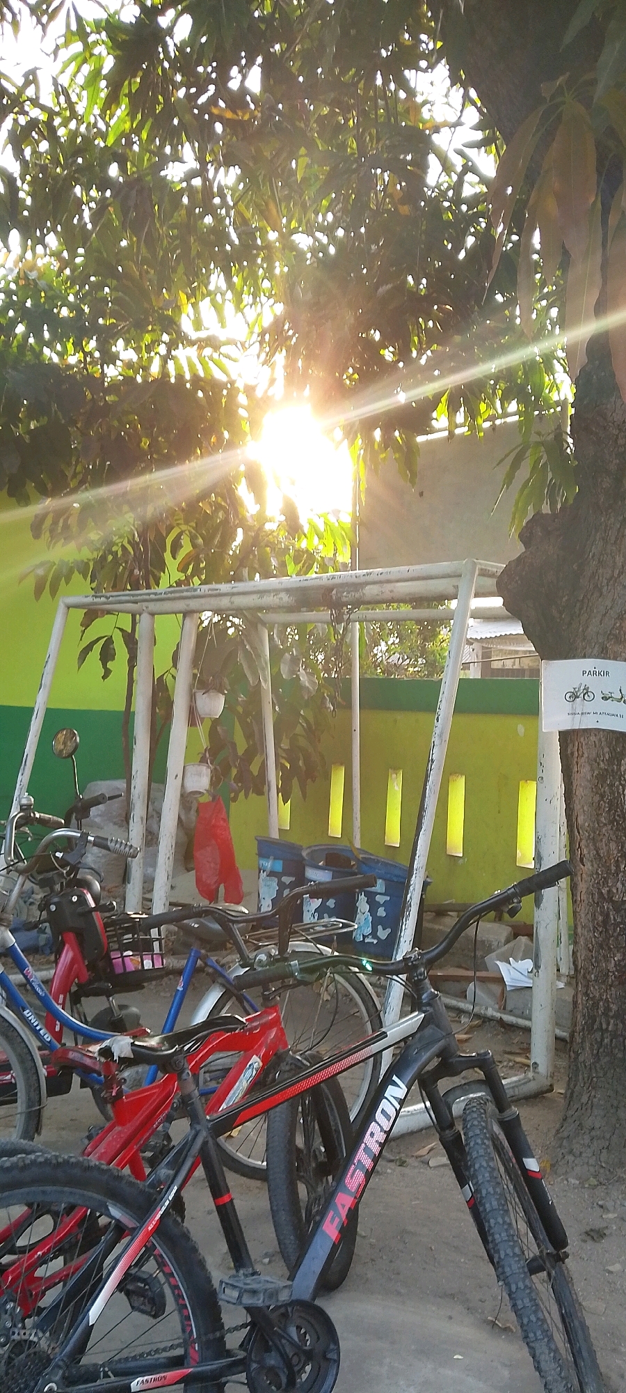 A group of bicycles are parked outdoors under a leafy tree in Satriajaya, Indonesia. The sun is shining brightly through the tree leaves, creating a lens flare effect. The bicycles are of various colors, including red and blue, and some appear to be mountain bikes. They are leaning against each other and a white metal frame structure. In the background, a bright green wall with yellow vertical accents is visible. A sign on a tree trunk reads "PARKIR" with images of a bicycle and a scooter. Two blue trash bins are also present. The ground is covered with dirt and gravel. There are no people visible in the image.