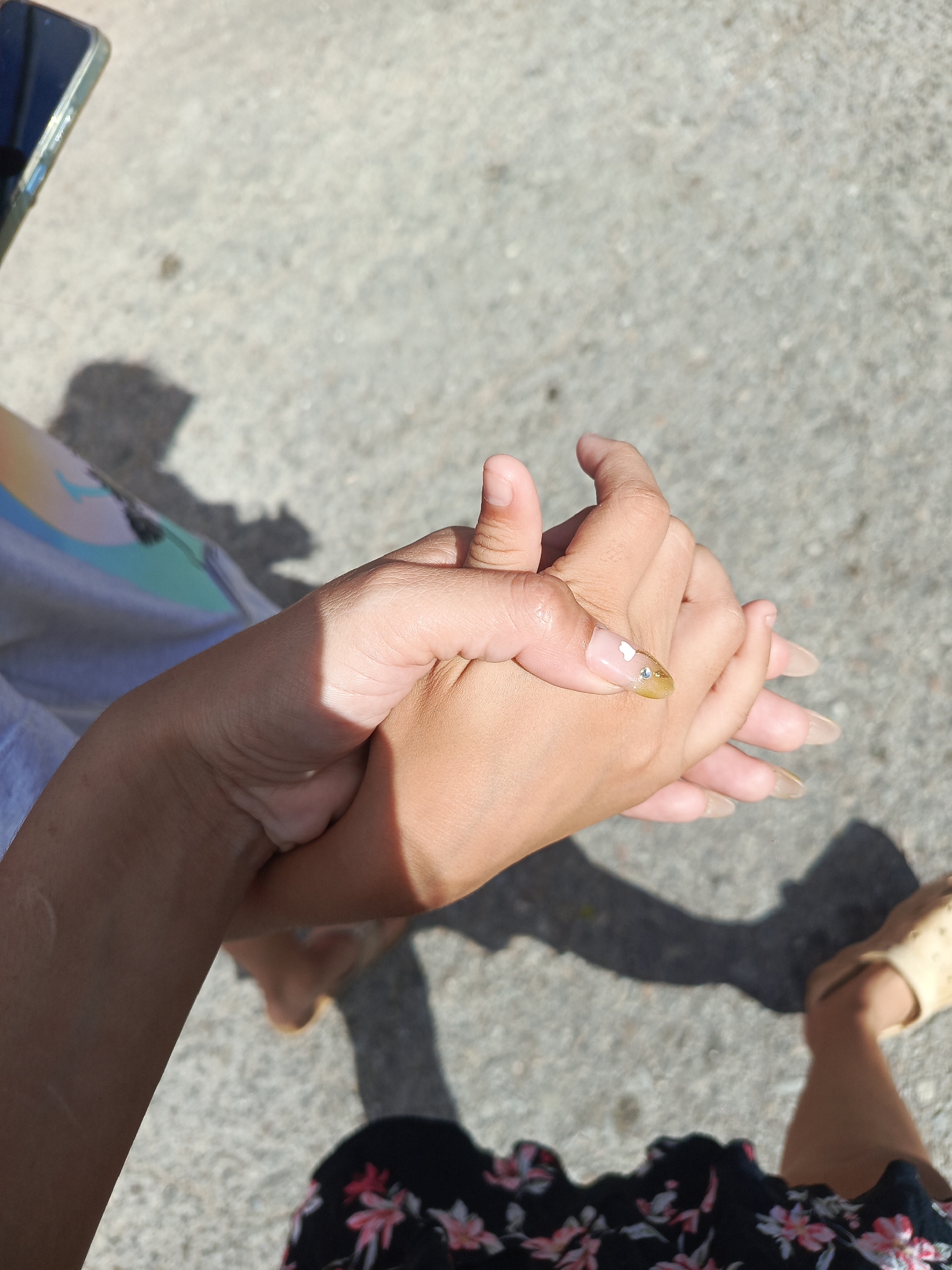 The image shows two hands clasped together, likely belonging to two individuals. One hand has long, oval-shaped nails with a nude color and one decorated nail with a gold tip and small white and rhinestone embellishments. The other hand has shorter, unpainted nails. The person holding the hands is wearing a floral-print dress or skirt. The footwear appears to be a pair of light-colored sandals. The setting appears to be outdoors on a paved surface, possibly concrete or asphalt. There are shadows cast on the ground, suggesting it is a sunny day. Based on the prompt's mention of Juan Griego, Venezuela, it can be inferred that the image was taken in that location.