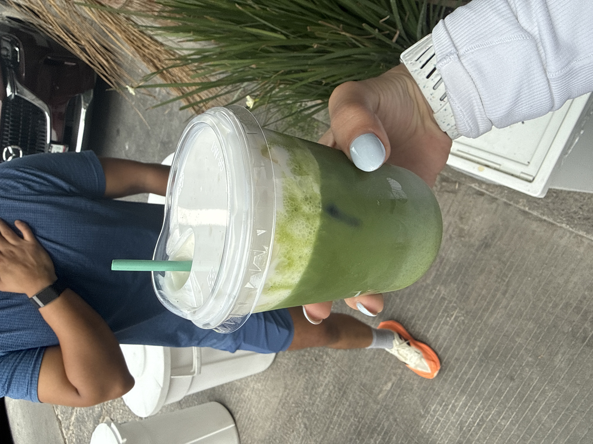 A person is holding a clear plastic cup with a green and white beverage, likely a matcha latte, outdoors. The person holding the cup has light blue nail polish and wears a white long-sleeved shirt with a sporty watch. Another person, wearing a blue shirt, shorts, socks, and orange running shoes, is partially visible. The background includes green foliage, a black car with a Mazda logo, and two white cylindrical objects, possibly planters or trash cans. The scene appears to be in an urban setting. Location cannot be determined from the image alone.