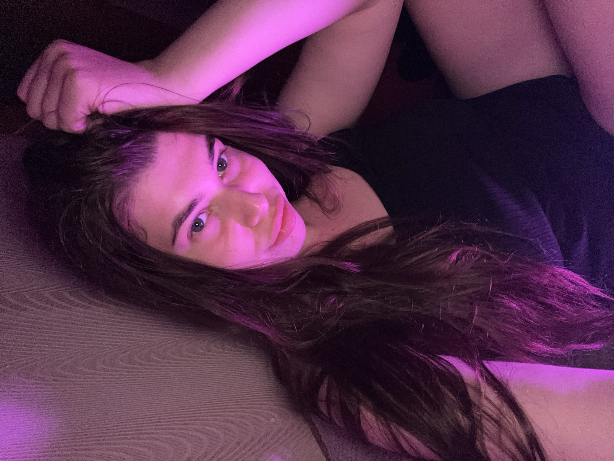 The image shows a young woman with long, dark hair, resting her head on her hand. She is wearing a black tank top. The scene is illuminated by purple and pink lighting, creating a vibrant and moody atmosphere. The background appears to be a textured wall, possibly wallpapered, with a subtle pattern. The overall impression is of a selfie taken in a dimly lit room. The location in the city of Мещёры, Russia, is not discernible from the visual elements in the image.