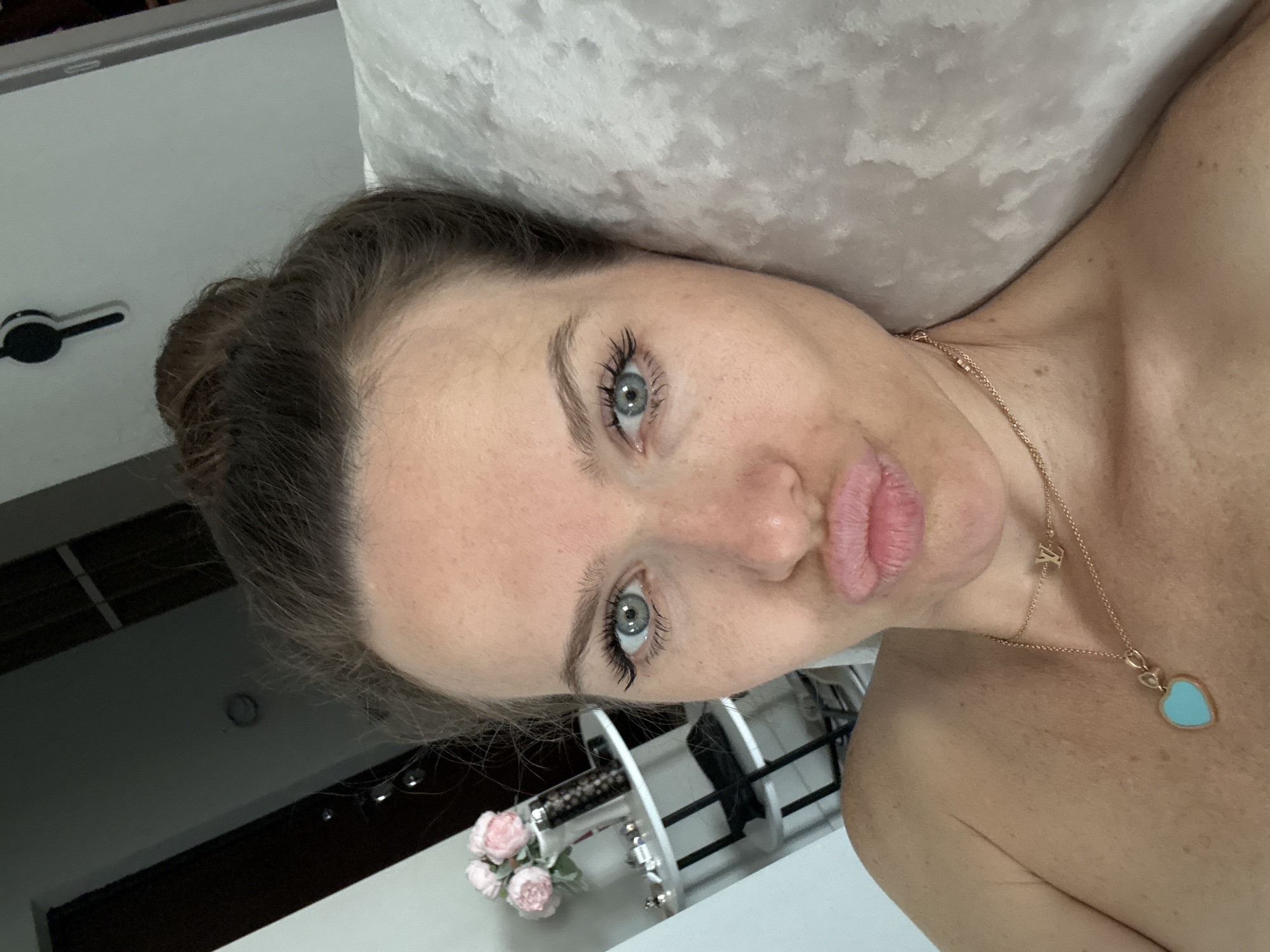 The image is a close-up portrait of a woman, rotated 90 degrees counter-clockwise, capturing her face and upper torso. She has fair skin, prominent light blue eyes, and dark brown hair pulled back from her face. Her eyelashes appear long and volumized, and her eyebrows are well-groomed. The woman is making a playful "duck face" expression with her full, pink lips. She is bare-shouldered, wearing a delicate gold chain necklace that features two small, stylized golden letters, "LV," near her collarbone, and a larger heart-shaped pendant crafted from a light blue material.
The background on the upper side of the image (when oriented correctly) consists of a soft, light-colored, textured fabric, resembling a pillow or blanket. On the lower side, a plain white wall is visible, featuring a black circular handle or knob. Near her shoulder, a decorative tiered stand, white with black accents, holds a small cluster of artificial pink roses. Other parts of the background show dark linear elements, possibly part of furniture or ceiling details. The setting appears to be indoors, likely a bedroom or private room, conveying a casual and intimate atmosphere. This media file was taken in Dolgoprudny, Russia.