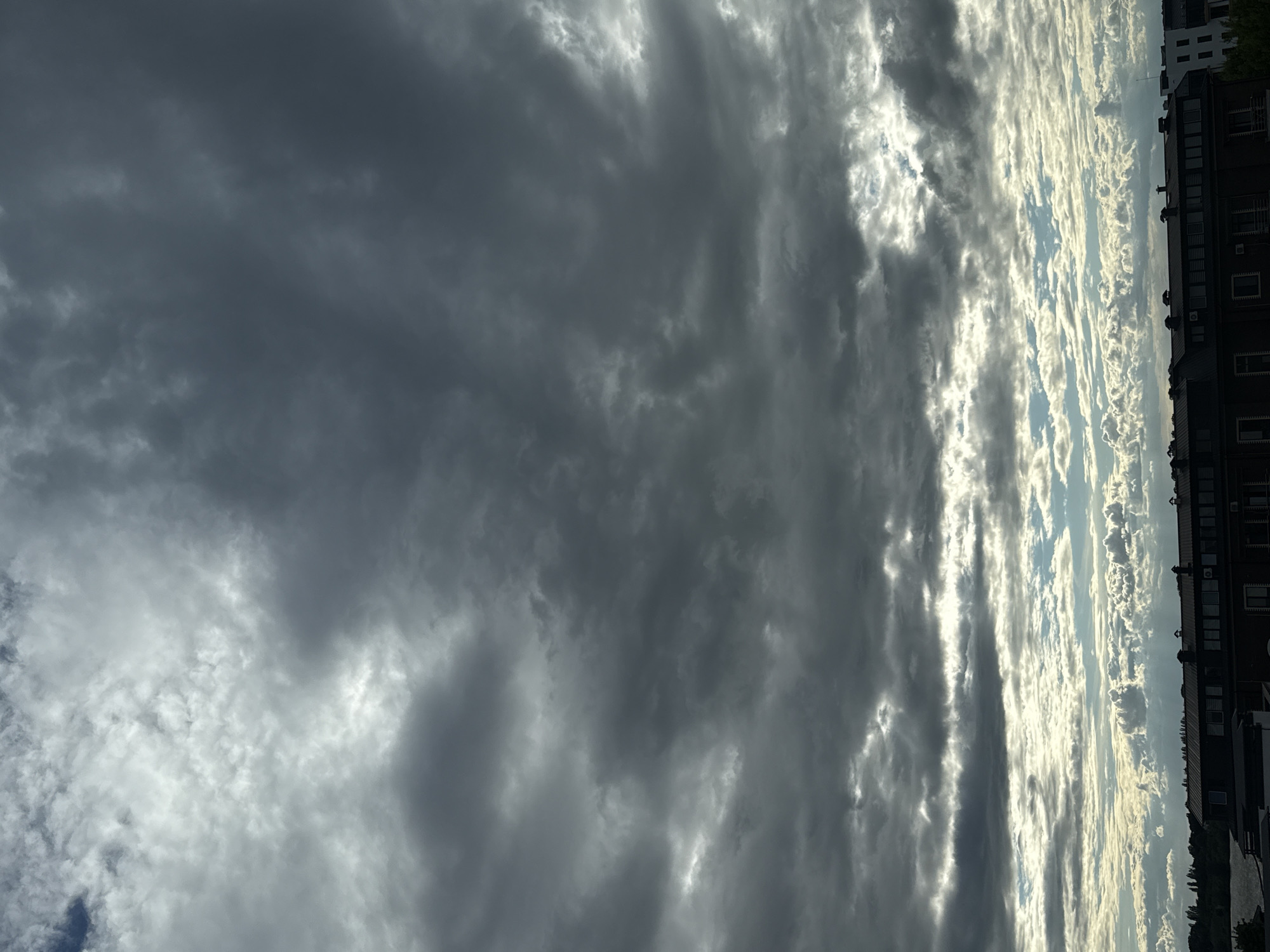 A dramatic, overcast sky dominates the image, with thick, dark clouds filling the upper two-thirds. The clouds are textured and varied, with lighter patches suggesting the presence of sunlight trying to break through. Below the heavy cloud cover, a layer of thinner, brighter clouds stretches across the horizon. These clouds are illuminated from behind, creating a luminous effect and revealing glimpses of blue sky.
The bottom portion of the image shows a silhouetted cityscape. A long, dark building with multiple windows and several visible vents or chimneys stretches across the frame. To the right, a white building with visible windows is partially visible. In the distance, a line of trees forms the edge of the landscape, with a body of water, possibly a river or lake, visible to the left of the trees. The overall impression is of a city under a moody, dynamic sky.