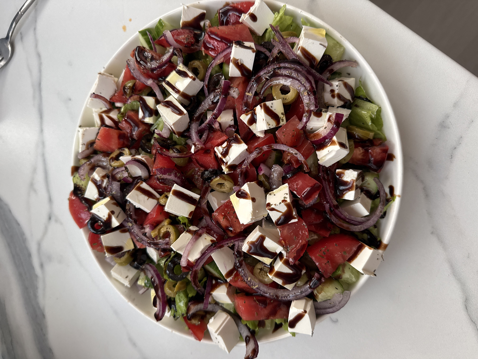 A close-up, top-down shot displays a generously portioned Greek salad, artfully arranged in a wide, white ceramic bowl. The salad is a vibrant medley of chopped tomatoes, cubed feta cheese, thinly sliced red onions, green and black olives, and leafy green lettuce. A dark, glossy dressing, likely balsamic glaze, is drizzled liberally over the ingredients, adding a rich sheen. The salad rests on a white, marble-patterned countertop. The composition is tightly framed, focusing exclusively on the food and its immediate surroundings. There are no people or actions depicted. The environment appears to be indoors, likely a kitchen or dining area. The image does not contain any information that would allow for a determination of its location in Sochi, Russia.