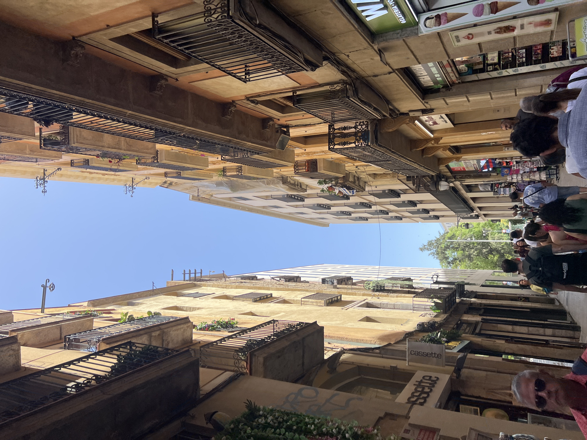 This media file captures a street scene in Barcelona, Spain, with buildings on either side of a narrow street bathed in bright daylight. The perspective is unusual, as the image is rotated 90 degrees counter-clockwise, making the buildings appear to stretch upwards into the sky.
The buildings are multi-story structures with traditional architecture, featuring balconies with wrought-iron railings. Some balconies have planters with greenery and flowers. The upper floors of the buildings are a warm, ochre color, while the lower levels vary in color and design, with some appearing to be shops or businesses.
Several people are visible in the image, walking along the street. They are casually dressed, suggesting a pleasant day for outdoor activity. The mood appears relaxed and everyday.
Notable details include signs on the buildings. One sign reads "cassette" with a minimalist design, and another below it also appears to advertise a similar business with stylized lettering. Further up the street, a sign with images of ice cream cones suggests a gelateria. The clear blue sky indicates good weather. The overall impression is of a bustling yet charming European street.