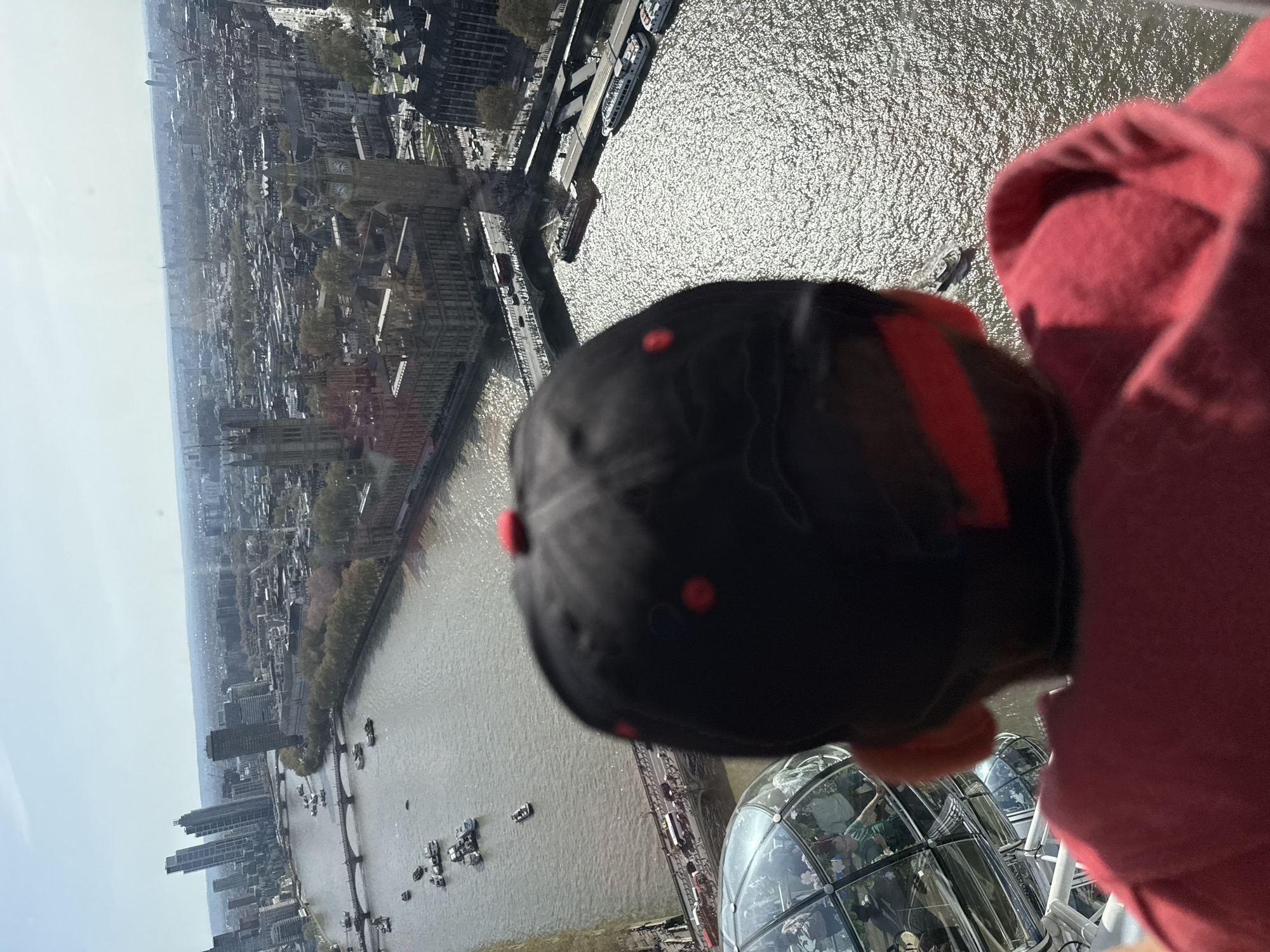 The image shows a view from inside the London Eye, looking out over the city of London, United Kingdom. The River Thames is visible with various boats and bridges crossing it. Notable buildings in the cityscape include the Houses of Parliament and Big Ben. A person wearing a red hoodie and a black baseball cap with red accents is in the foreground, partially obscuring the view. The interior of the London Eye capsule is also visible, with other people inside. The image is taken from a high vantage point, giving a panoramic perspective of the city.