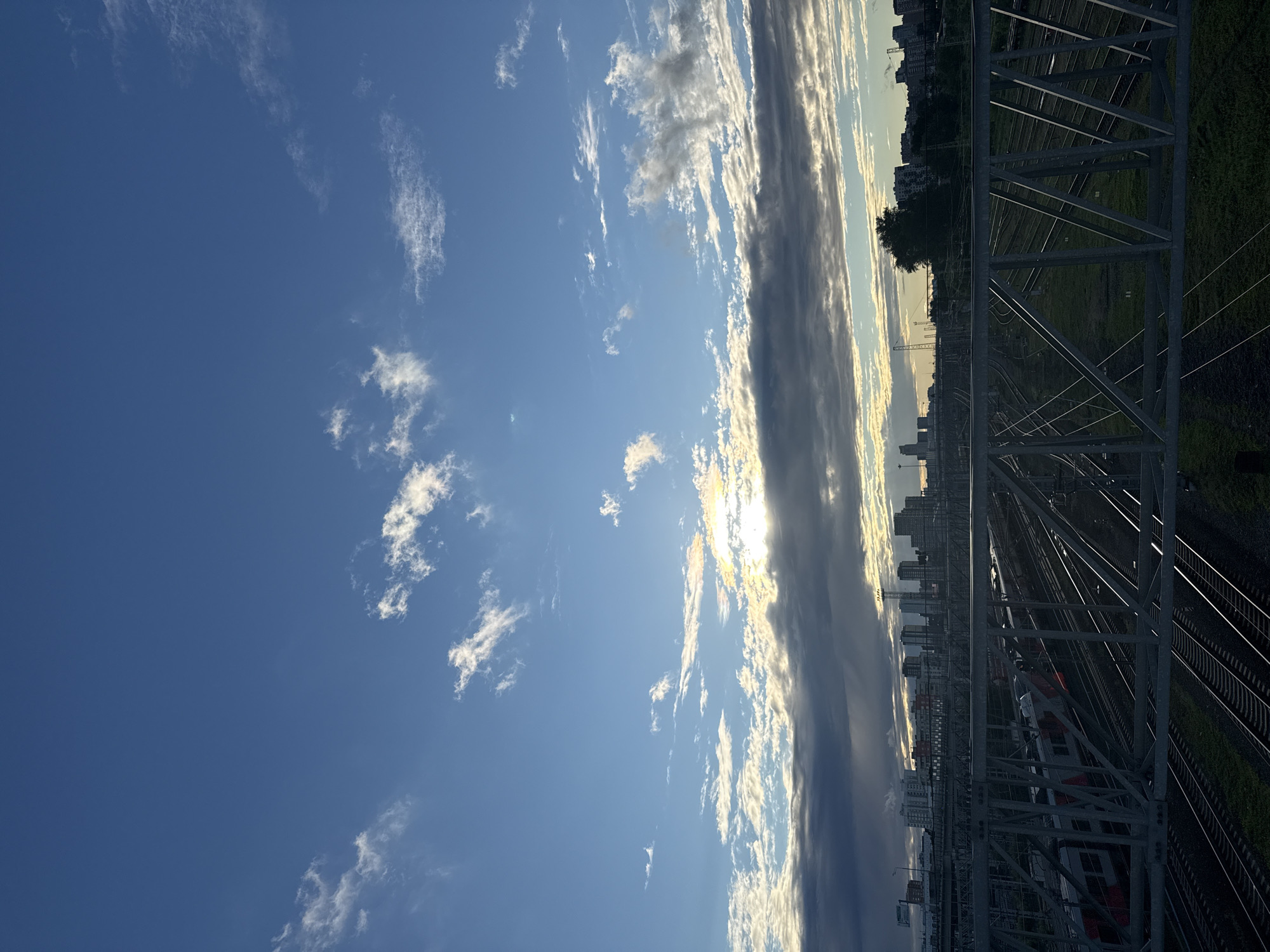 This is a vertically oriented photograph of a sky filled with clouds, with a train track structure dominating the right side. The sky is predominantly a clear, bright blue with scattered white and grey clouds. Some of the clouds are thin and wispy, while others are denser and streaky, suggesting movement and texture. The sun is partially obscured by a band of clouds, creating a bright glare and illuminating the edges of the clouds with a golden hue.
Below the sky, the right side of the image features a large, metallic, triangular-truss structure, likely part of a railway overpass or bridge. Through the gaps in this structure, multiple sets of train tracks are visible, stretching into the distance. Green grass borders the tracks. In the background, a city skyline is visible, with numerous multi-story buildings. A single tree with dark foliage stands out against the buildings. The overall lighting suggests it is either early morning or late afternoon, with the low angle of the sun casting long shadows and creating a warm, atmospheric light. A red and white train is partially visible on the tracks. The scene appears to be a railway yard or station in an urban environment, possibly Moscow, Russia, given the building architecture. There is no visible text.
