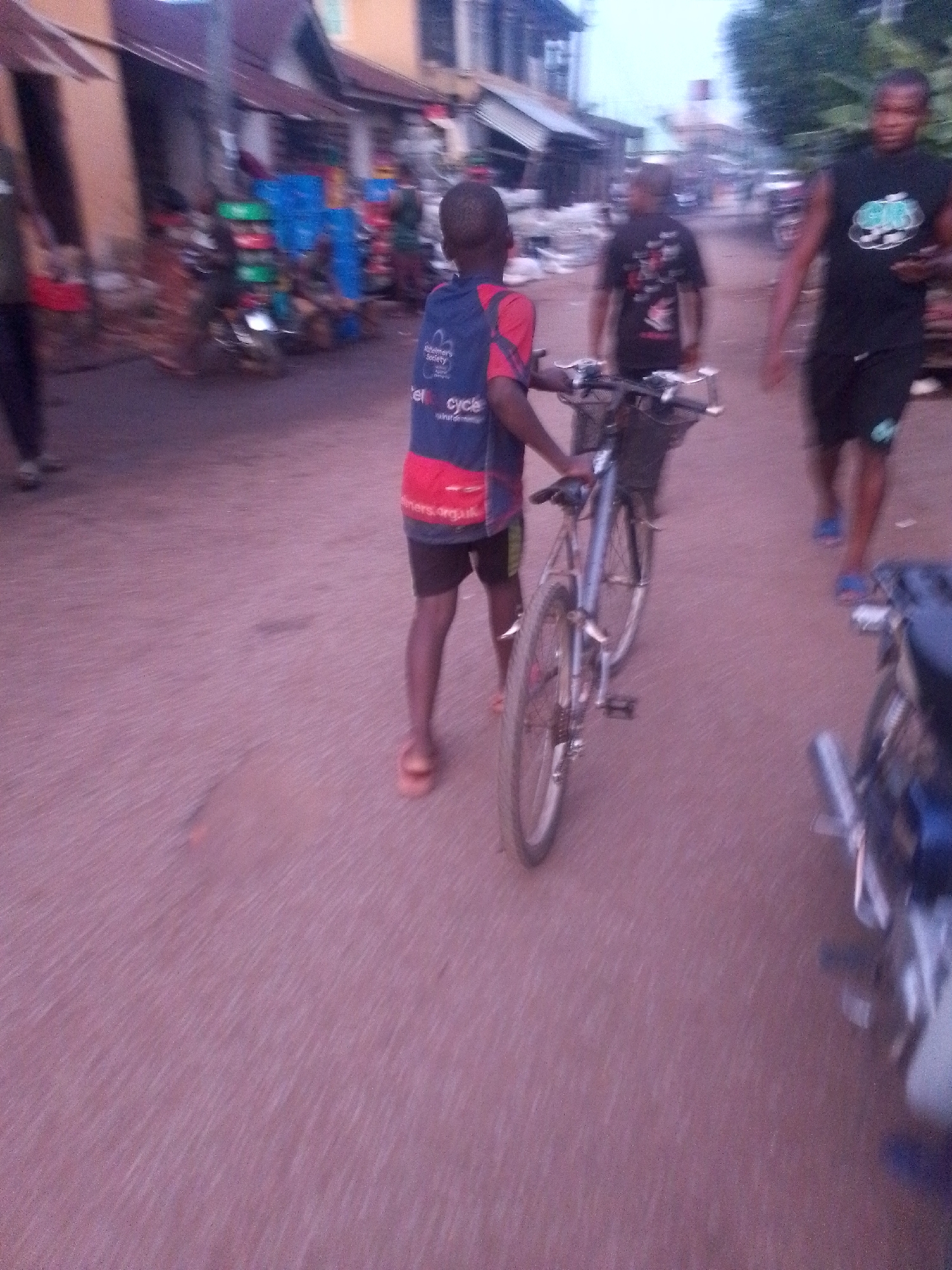 The image depicts a street scene in Orlu, Nigeria. A young boy is walking with a bicycle. Two other individuals are seen walking on the street in the background. There are buildings visible along the street, some with stalls or displays outside. A motorcycle is parked on the side of the road. The road surface appears to be reddish.