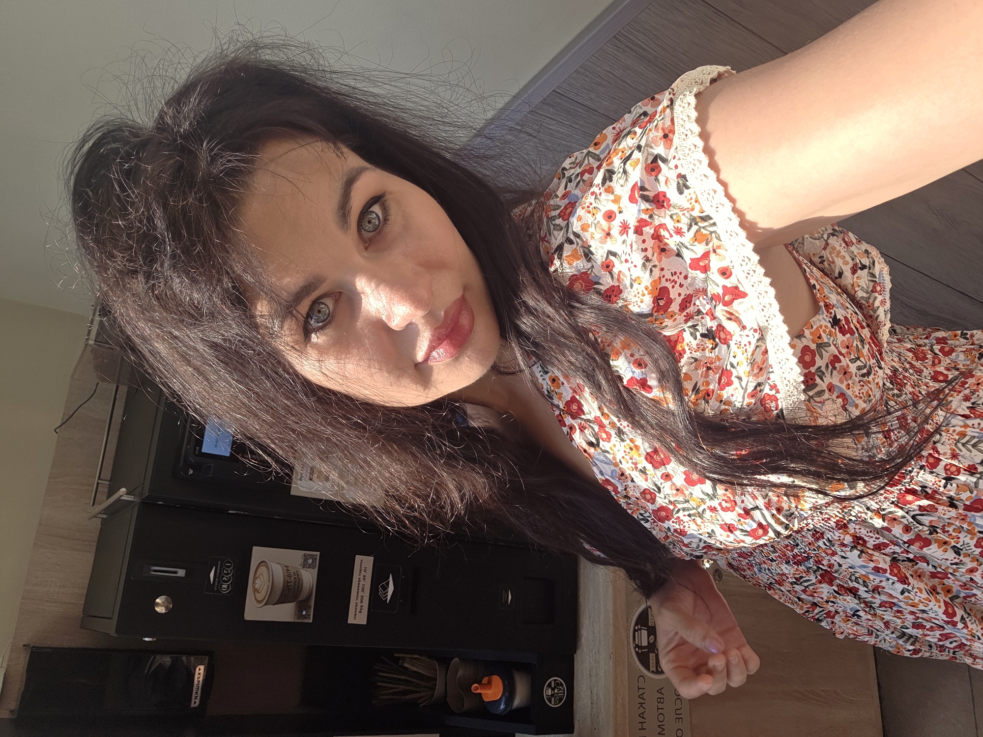 In an indoor setting that appears to be a cafe or break room, a woman with long, dark hair and blue eyes is taking a selfie. She is wearing a floral print dress with short sleeves and a lace trim. She is positioned to the right of the frame, with her face tilted slightly towards the camera. Behind her, on the left side of the image, is a commercial coffee machine. The machine is black and has various buttons and a screen. A sticker on the machine features an image of a latte with latte art. To the left of the coffee machine is a dispenser containing stir sticks and sugar packets. The background consists of wood-paneled walls and a wooden floor. The lighting suggests natural light is present, casting shadows on the floor. The location is identified as Sochi, Russia.
