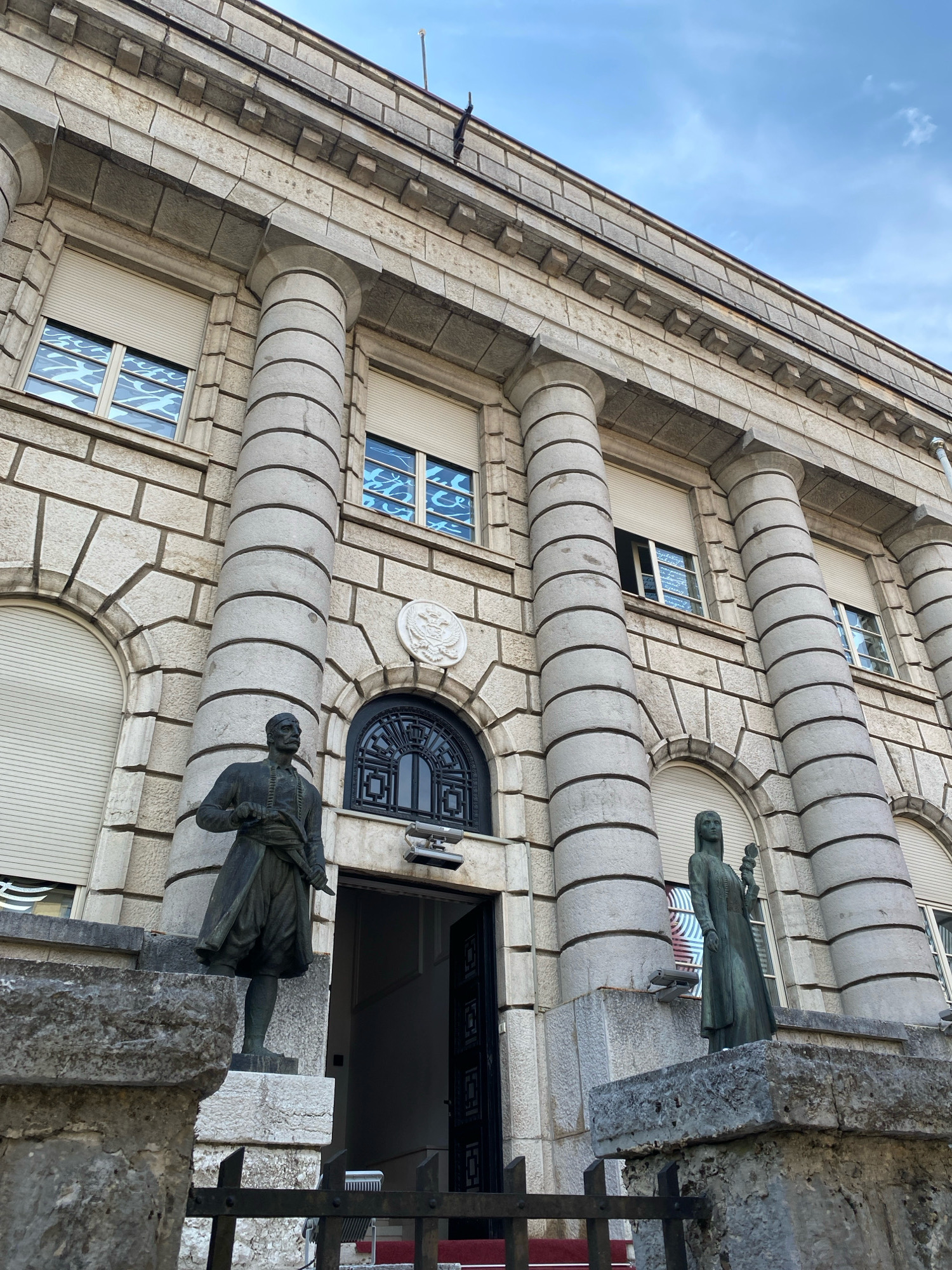 The image shows the front facade of a stone building, likely a government institution, in Cetinje, Montenegro. Two bronze statues stand on pedestals in front of the building. The statue on the left depicts a man in traditional clothing holding a sword or dagger, while the statue on the right shows a woman holding a flower. The building features large, fluted columns, arched windows with rolling shutters, and a prominent arched doorway. Above the doorway is a circular emblem. The sky above is blue with some scattered clouds.