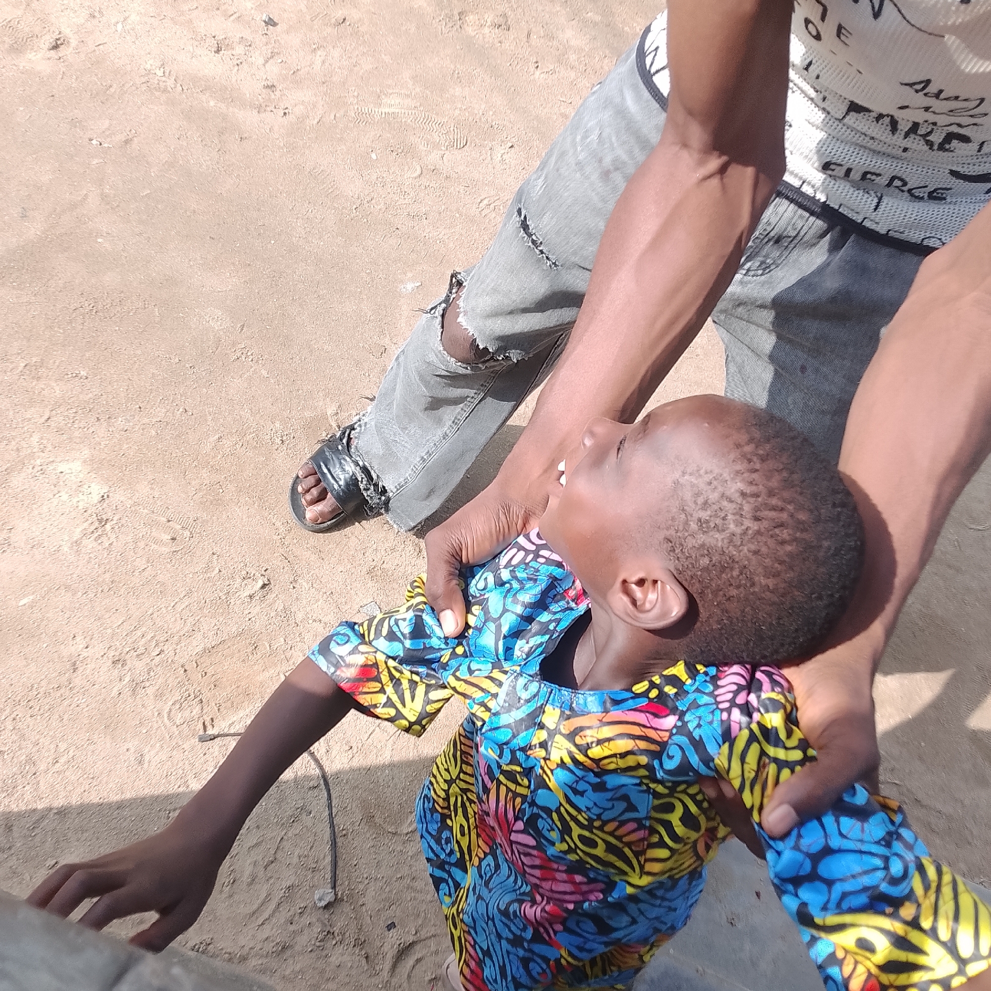The image depicts a child wearing a colorful, patterned shirt, seemingly being held or supported by an adult. The child's head is tilted back, and their mouth is open, suggesting they might be laughing or speaking. The adult's arms are visible, one of which is supporting the child. The adult is wearing ripped jeans and a white t-shirt with black text. The scene appears to be outdoors, on sandy ground, indicative of a potentially rural or less developed urban environment. The lighting suggests a sunny day. The overall impression is one of playful interaction. This scene could plausibly be located in a neighborhood within Ado, Nigeria, given the appearance of the subjects and the environment, which is consistent with many areas in Nigerian cities.