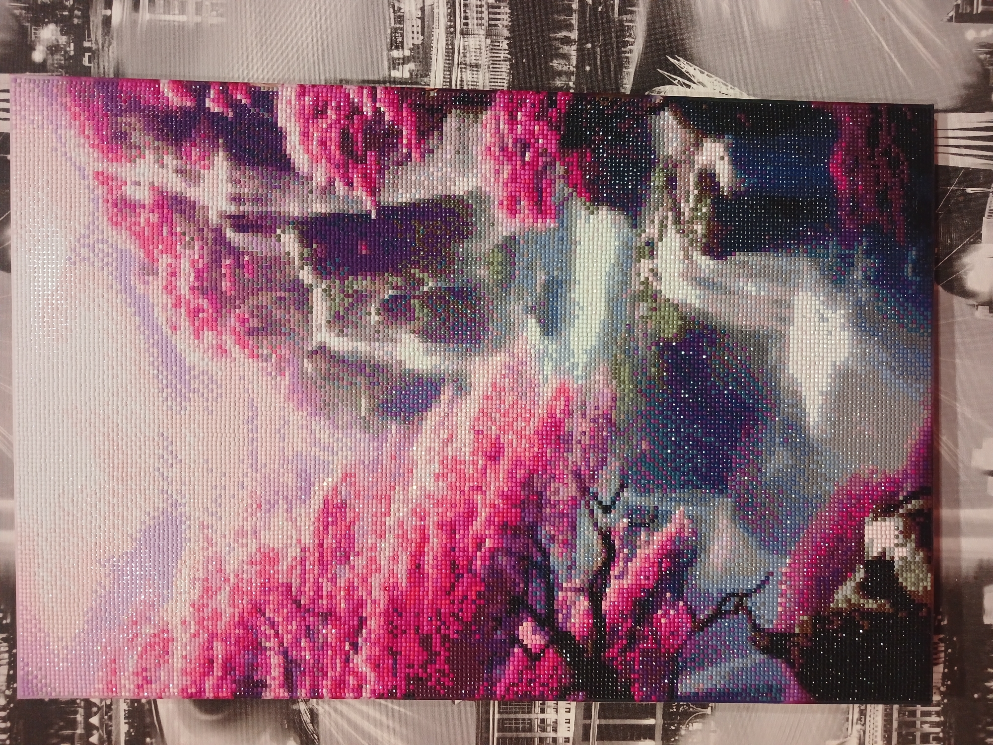 The image shows a rectangular diamond painting with an impressionistic view of cherry blossoms in full bloom against a blurred urban backdrop. The foreground features intense pink and purple hues forming the blossoms and branches of the cherry trees. A bright, white-to-blue gradient occupies the center, suggesting a body of water or sky. The background, though indistinct, hints at buildings and cityscape elements rendered in soft, muted colors. The painting is displayed on a surface with a monochrome cityscape pattern. There are no people visible in the scene. The overall impression is of a stylized, decorative artwork depicting a serene natural scene contrasted with a modern city. The location of Noviy, Russia is not evident from the image.