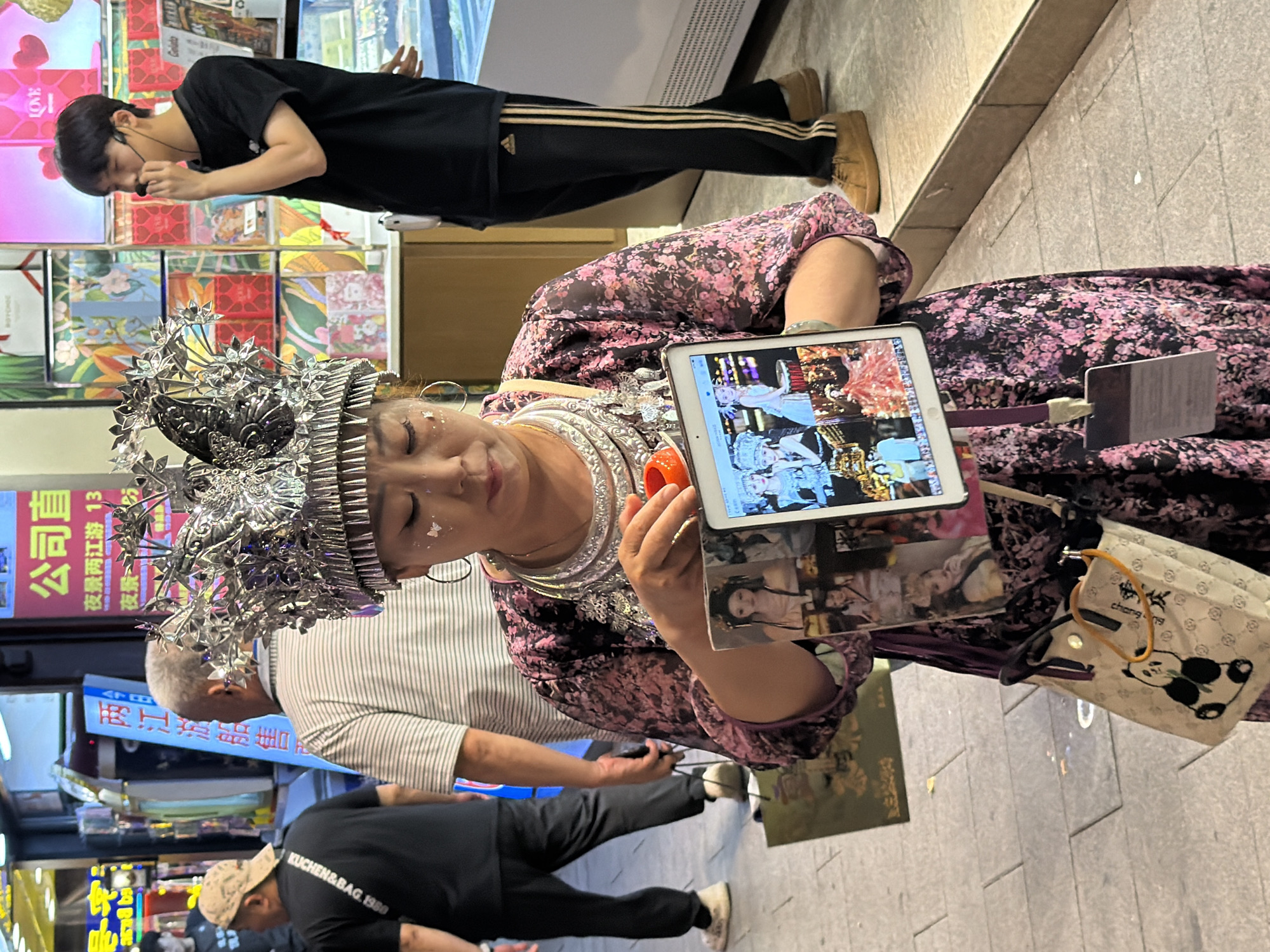 A group of people are gathered in what appears to be a bustling street market or shopping area in Yuzhong District, China. In the foreground, a woman adorned in traditional ethnic attire, including a highly ornate silver headdress and a large silver necklace, is holding a tablet computer and a small, orange, round object. She wears a floral print dress. Her eyes are closed, and she appears to be deep in thought or enjoying a moment of calm amidst the activity.
Behind her, a man in a black t-shirt with distinctive stripes on his pants walks away from the camera. Further back, another man in a black t-shirt with text that reads "KUCHEN&BAG. 1980" and a baseball cap is partially visible, also moving away. In the background, there are displays of various items, possibly products or promotional materials, with some featuring vibrant colors and floral designs. Signs with Chinese characters are also visible, indicating the commercial nature of the setting. One sign displays the text "公司直" and "夜景两江游 13", suggesting tourism or local attractions. A panda-themed handbag hangs from a strap on the woman's shoulder. The overall atmosphere seems to be that of a lively daytime scene, with the presence of people and commercial activity.