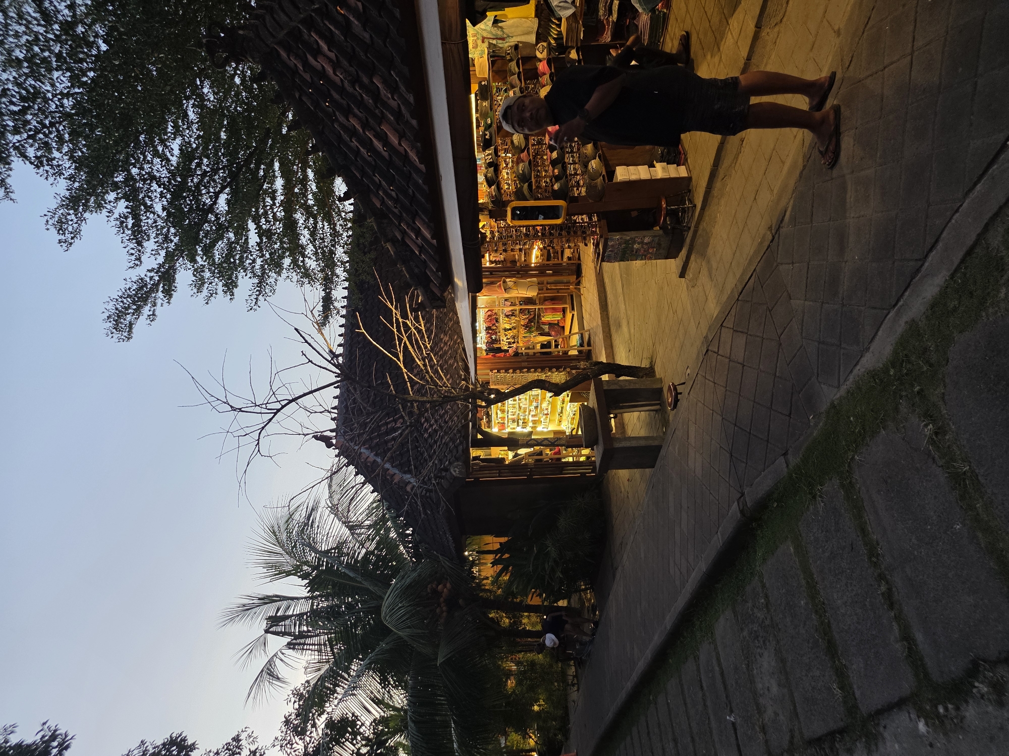 The image depicts a street scene in North Kuta, Indonesia at dusk or night. A man wearing a black shirt, shorts, and a white cap is standing in front of a brightly lit shop with a traditional tiled roof. The shop's interior displays various goods on shelves. To the left of the shop, trees, including a palm tree, are visible against a dusky sky. The foreground shows a paved walkway and a low wall with grass growing in the gaps.