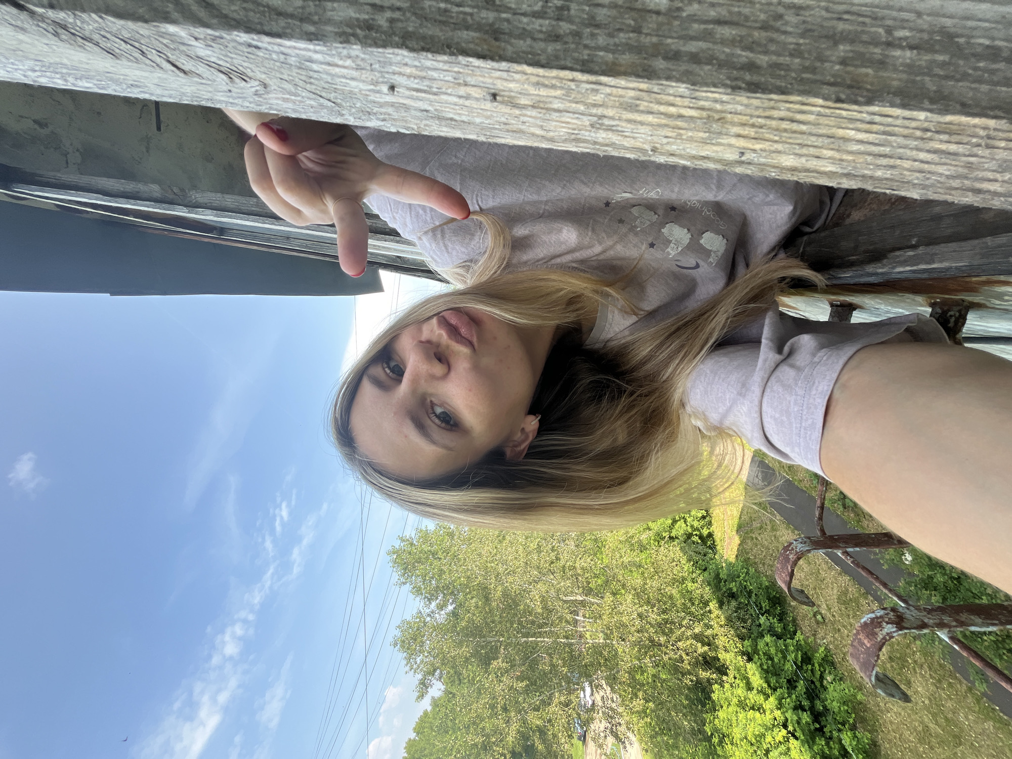 In a selfie taken from a balcony in Chaikovsky, Russia, a young Caucasian woman with blonde hair and light skin is leaning out from behind a weathered wooden structure. She is wearing a light purple t-shirt with a graphic print of llamas and the words "Goodnight, Sleep Tight." Her lips are pursed in a duck face gesture, and her right hand is extended, forming a peace sign. Behind her, a blue sky with scattered white clouds is visible, along with green trees and what appears to be a residential area with parked cars and power lines. The balcony itself is framed by rusty metal railings and the rough texture of the wooden beam. The overall impression is a casual, playful moment captured outdoors on a sunny day.