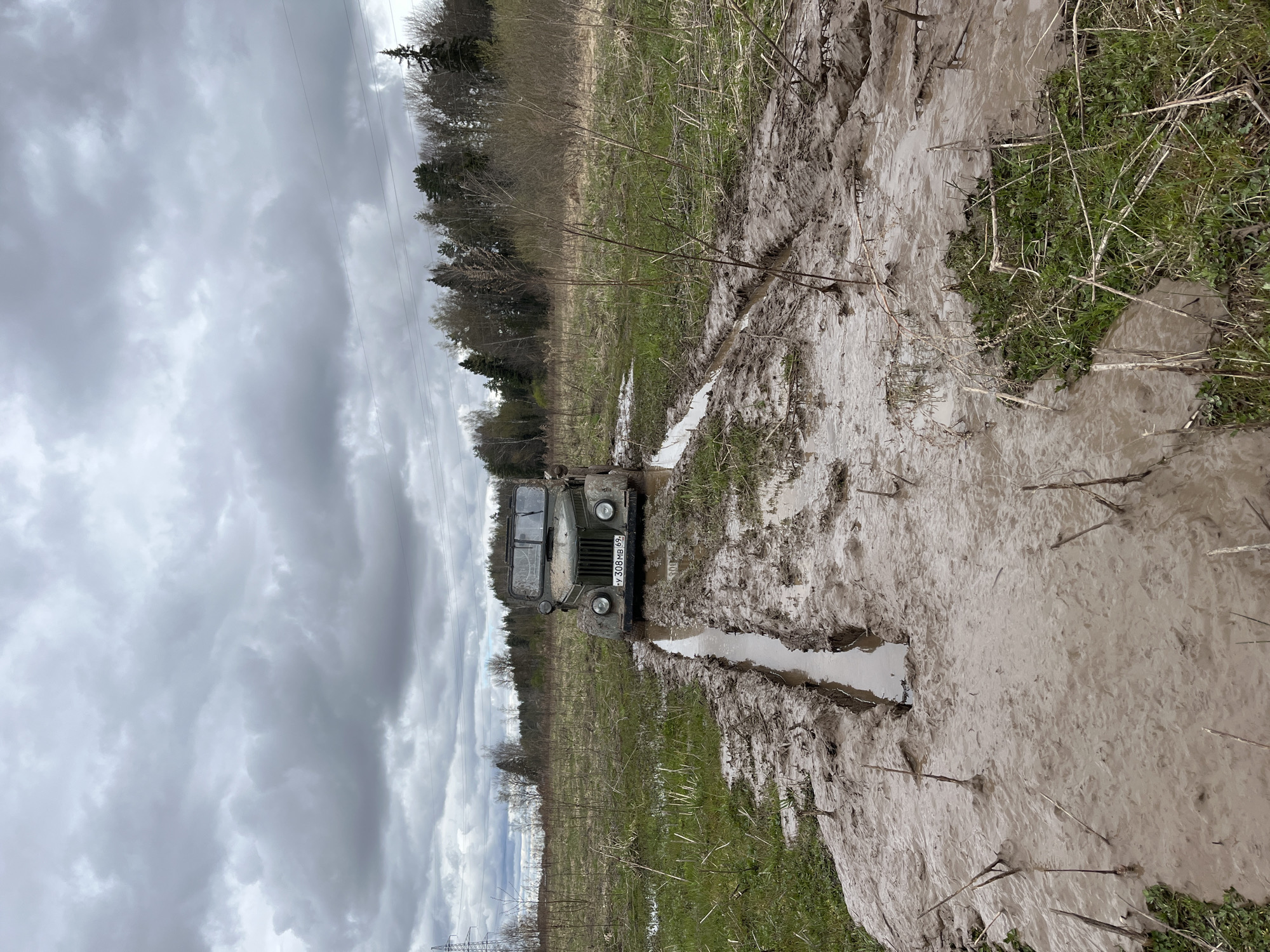 The image depicts a military-style truck driving through a muddy field. The field is located near a wooded area under a cloudy sky. Power lines are visible in the background. The truck is covered in mud, suggesting difficult driving conditions. The vehicle’s license plate appears to read "У 308 МВ 69". The exact location within Раздолье, Russia, cannot be determined from the image alone.