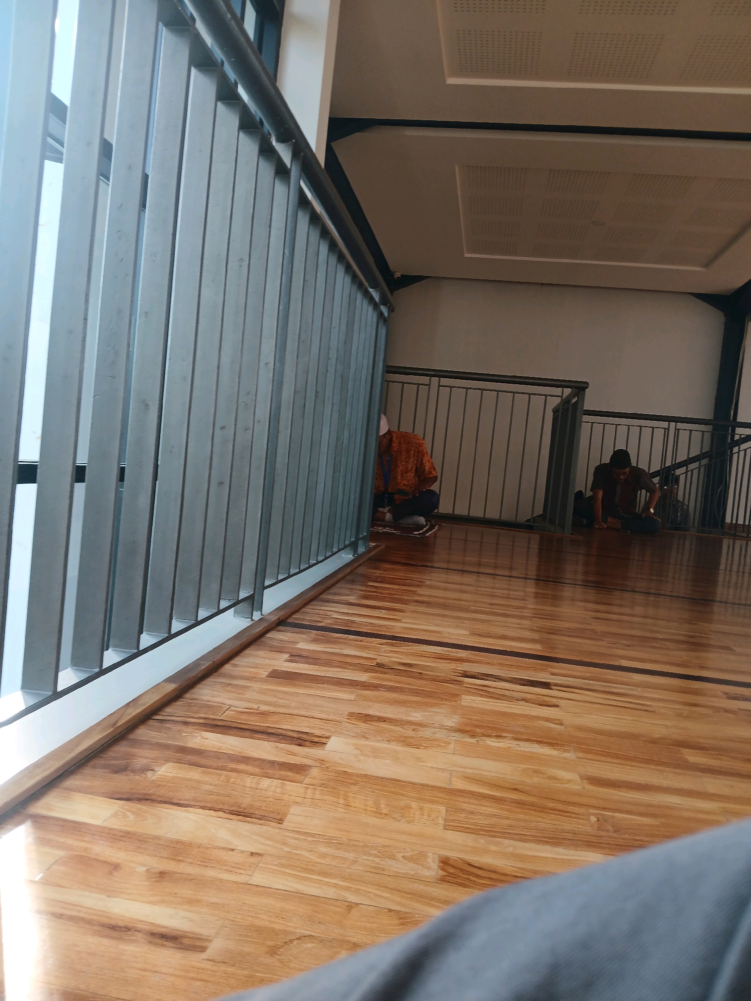 The image depicts an indoor scene with a polished wooden floor and metal railings. Two individuals are visible, both appearing to be in a seated or kneeling position on the floor. One person on the left wears an orange patterned shirt and is partially obscured by a railing. The other individual on the right is in a brown shirt and is seated on the floor. The environment suggests a public indoor space, possibly a hall or common area. The specific location in Arjasari, Indonesia, is not visually evident from the image itself.