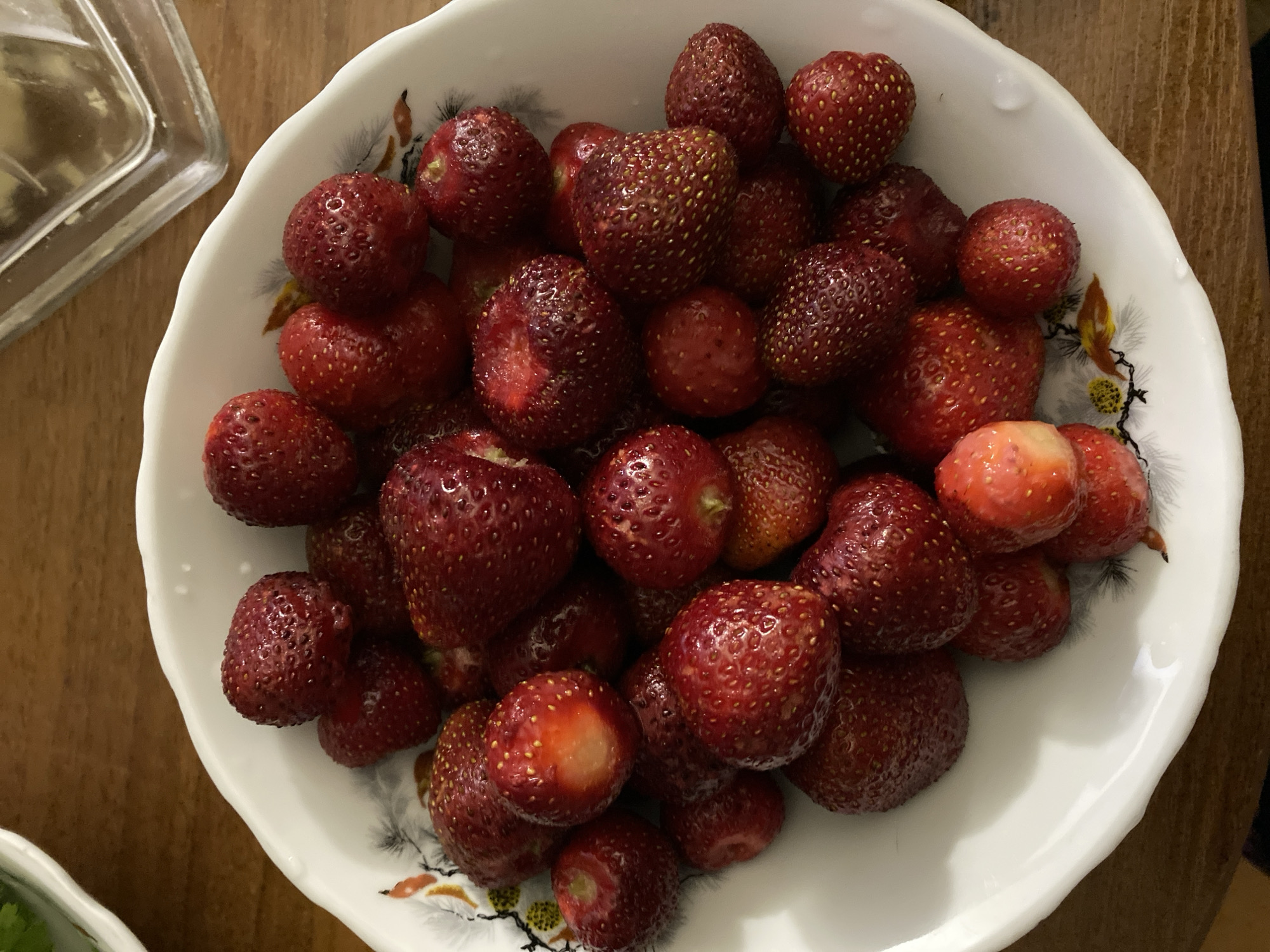 A close-up, overhead shot shows a bowl filled with ripe strawberries. The strawberries are various shades of red and are piled high in a white bowl with a decorative pattern of leaves and flowers along the rim. The bowl rests on a wooden surface. In the background, a clear glass object, possibly a container or a part of a dispenser, is visible. The lighting suggests an indoor setting. There are no people or animals in the image.