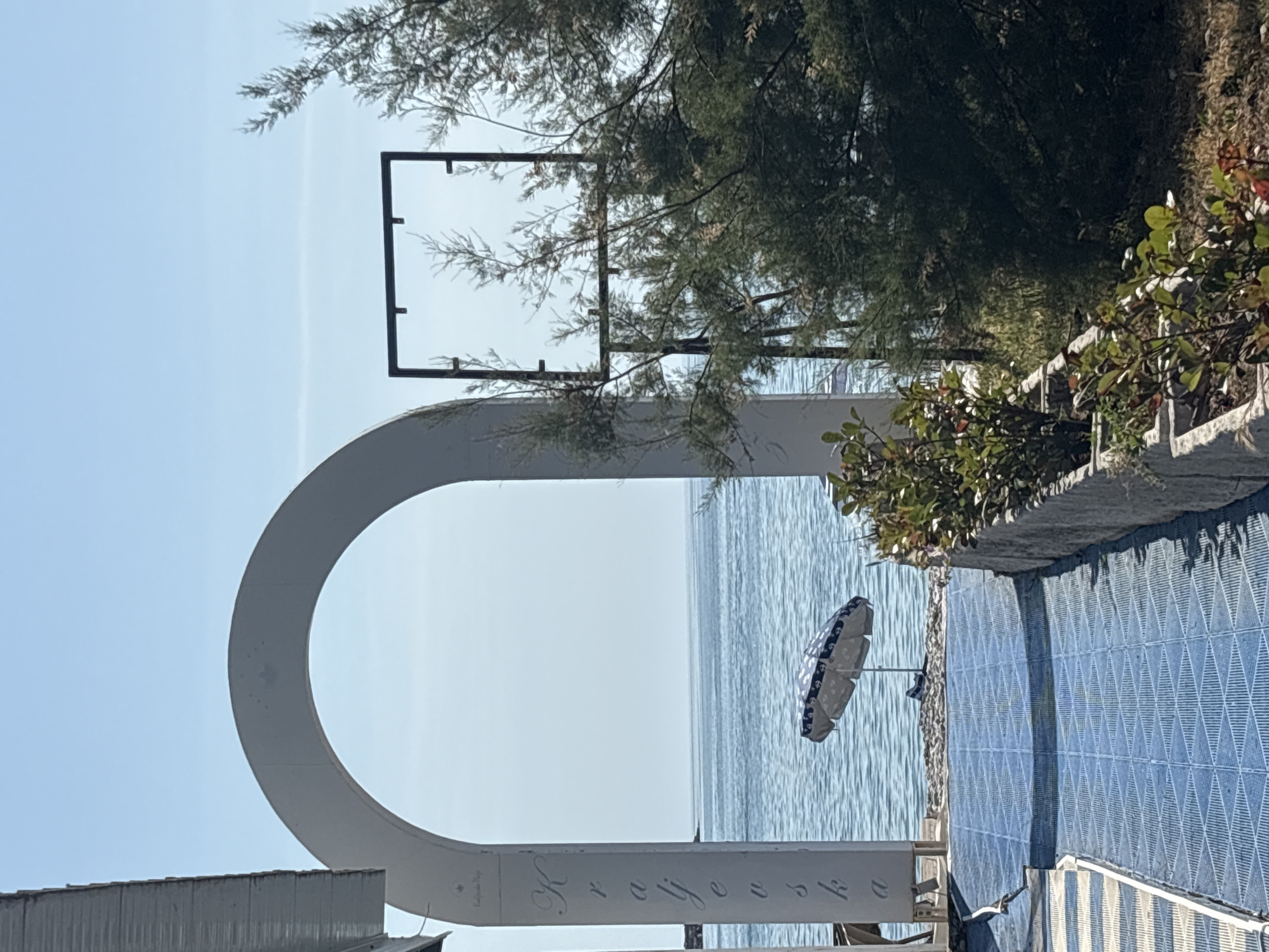 The image depicts a coastal scene in Bar, Montenegro, viewed through a white arched structure with the name "Kafić" written on it. A black rectangular metal frame sits above the arch. Beyond the structure, the Adriatic Sea stretches to the horizon under a clear, light blue sky. A single beach umbrella is visible on the shore. Vegetation, including trees and bushes, frames the right side of the view. A blue tiled structure is on the bottom right.