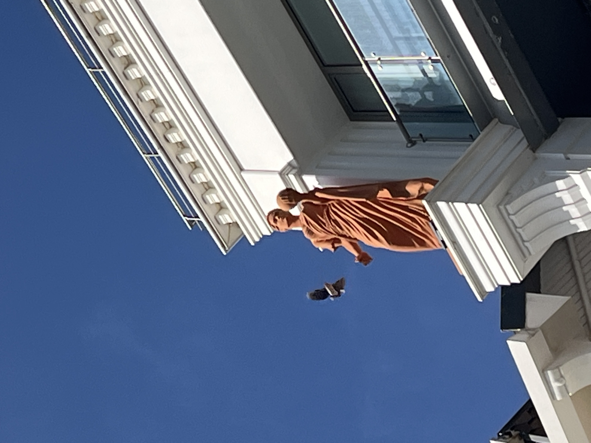 A low-angle shot captures a building in Alanya, Turkey, featuring a tan statue under a blue sky. A bird flies between the statue and the sky. The building has white architectural details.