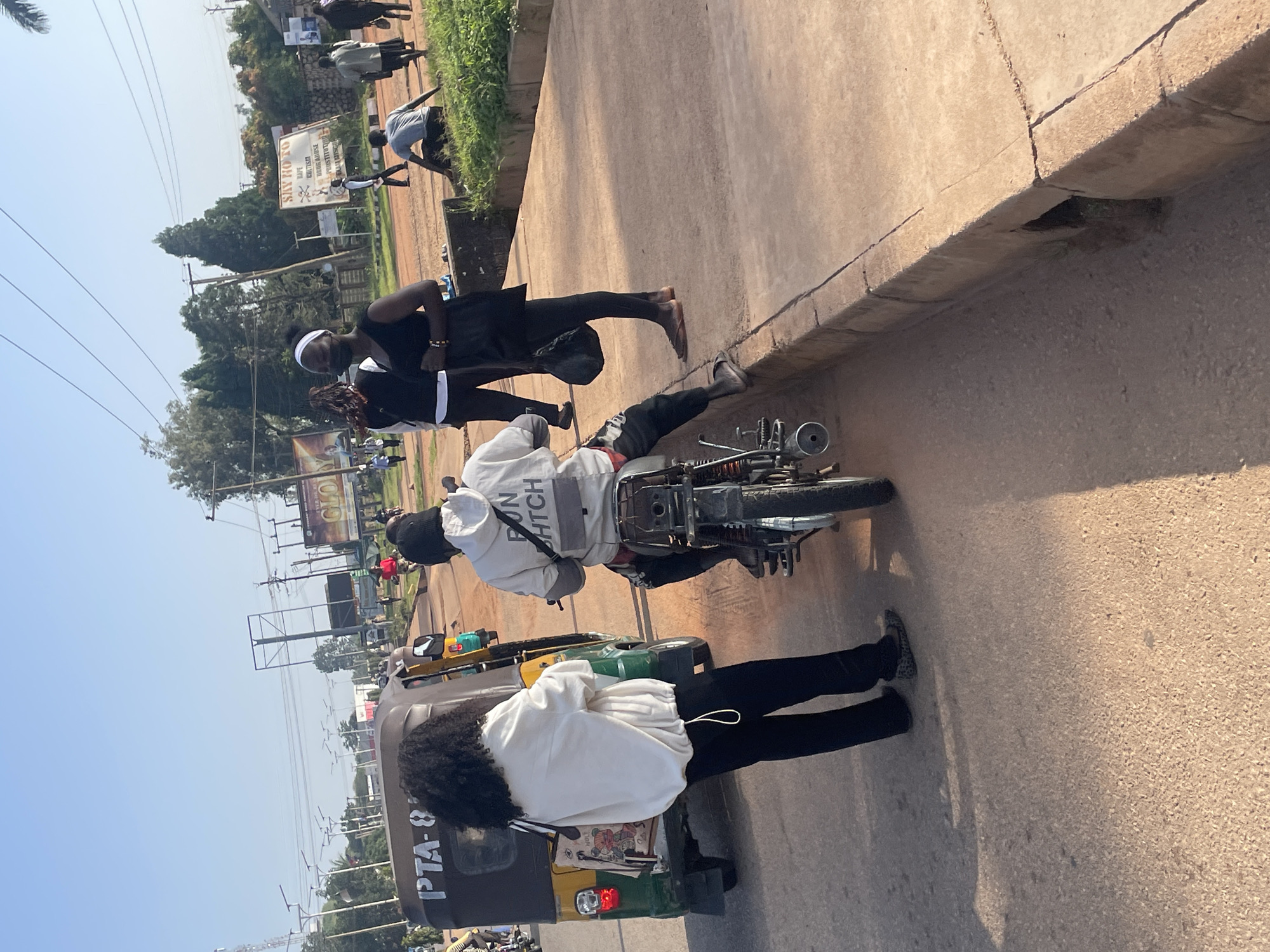 In Jos, Nigeria, on a sunny day, a man wearing a white hoodie with "DON'T HITCH" emblazoned on the back, rides a motorcycle. Beside him, a woman with dark, curly hair and wearing a white jacket and black pants stands next to a yellow and green auto-rickshaw with "PTA - 8" visible on its side. Another woman, wearing a black dress and a black face mask, stands on the sidewalk. Several other individuals are visible in the background, some walking and others on motorcycles. The scene takes place on a paved road with a dirt shoulder lined by grass and a concrete curb. Billboard advertisements are visible in the distance.