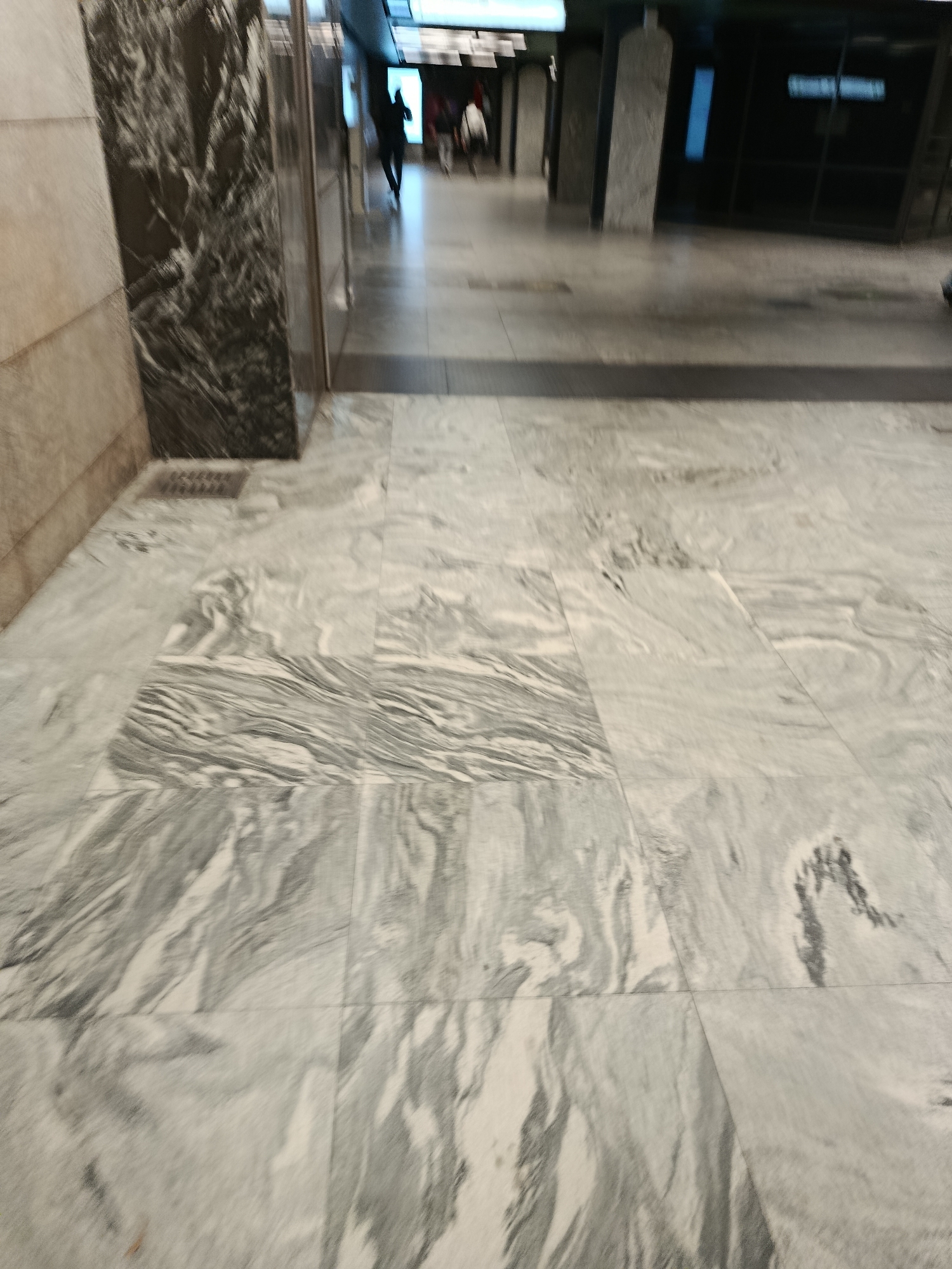 The image shows the marble floor of a public building, possibly a metro station or a shopping mall, in Moscow, Russia. The floor is made of large, polished marble tiles with a distinctive gray and white veined pattern. In the background, several people are walking in a hallway, their figures blurred due to motion. The lighting is bright, illuminating the reflective surfaces of the marble. The scene appears to be indoors, with architectural elements like pillars and glass partitions visible in the background.
