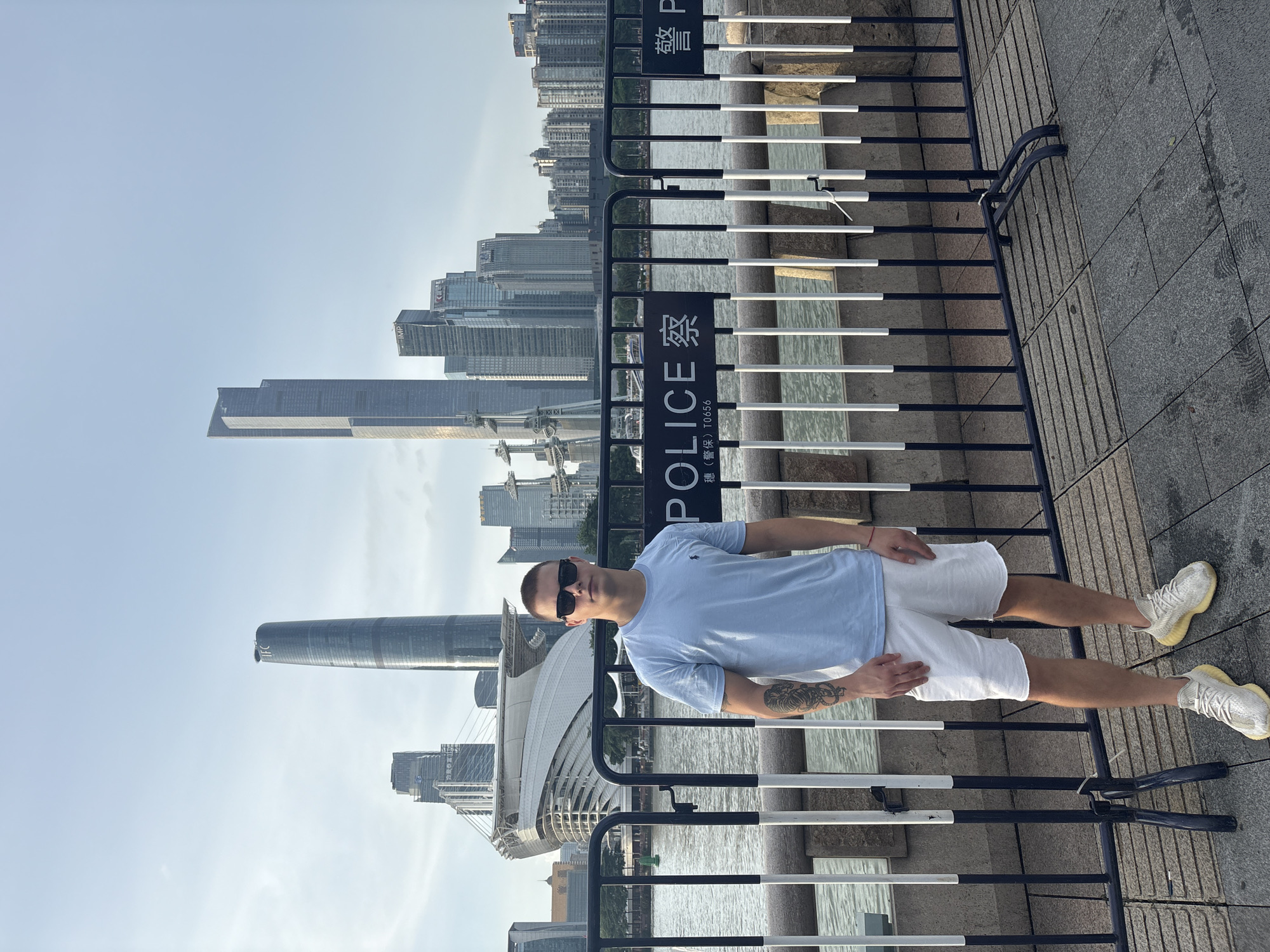 The image shows a young man standing outdoors in front of a fence with a cityscape in the background. The man is wearing a light blue t-shirt, white shorts, and sunglasses. He has a tattoo on his left arm. Behind him, a river or body of water separates the foreground from a densely populated urban area featuring numerous skyscrapers and modern buildings. A large, distinctive curved building with a white facade is also visible. The sky is clear and bright, suggesting it is daytime. The fence in the foreground has signs with text in both English and Chinese. One sign clearly reads "POLICE" vertically, and the Chinese characters above it translate to "Police". The overall impression is of a tourist or resident enjoying a view of a modern city. The scene appears to be in Haizhu District, China.