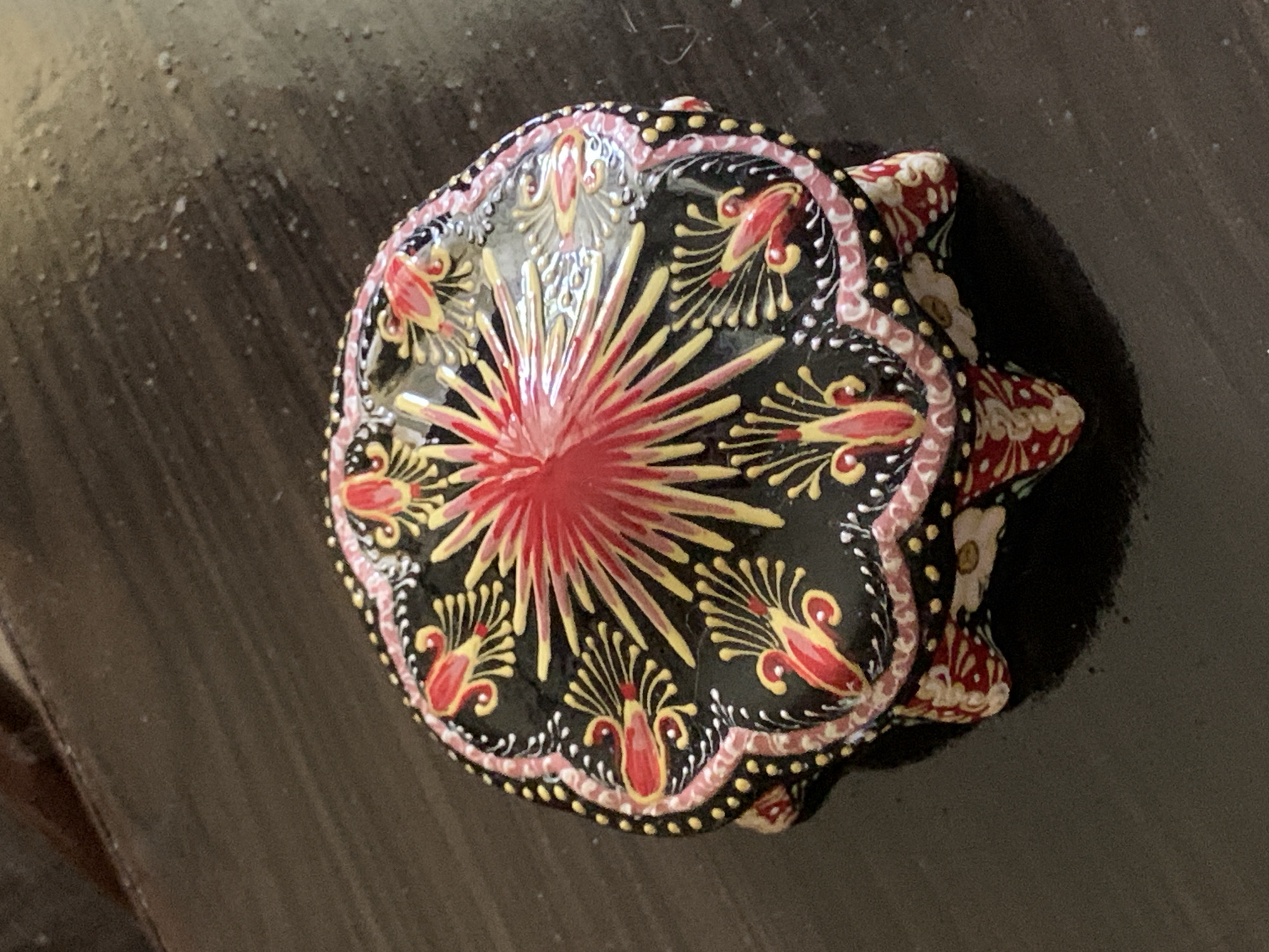 A close-up, overhead shot shows a decorative, round, lidded trinket box with intricate painting. The box has a black base adorned with floral and geometric patterns in red, yellow, and white. The lid features a radiating starburst design in red and yellow at its center, surrounded by floral motifs. The box rests on a dark, textured surface, likely a wooden table. The lighting suggests an indoor setting. This object is a handicraft, possibly a souvenir, and could be found in a market or home in Tashkent, Uzbekistan.