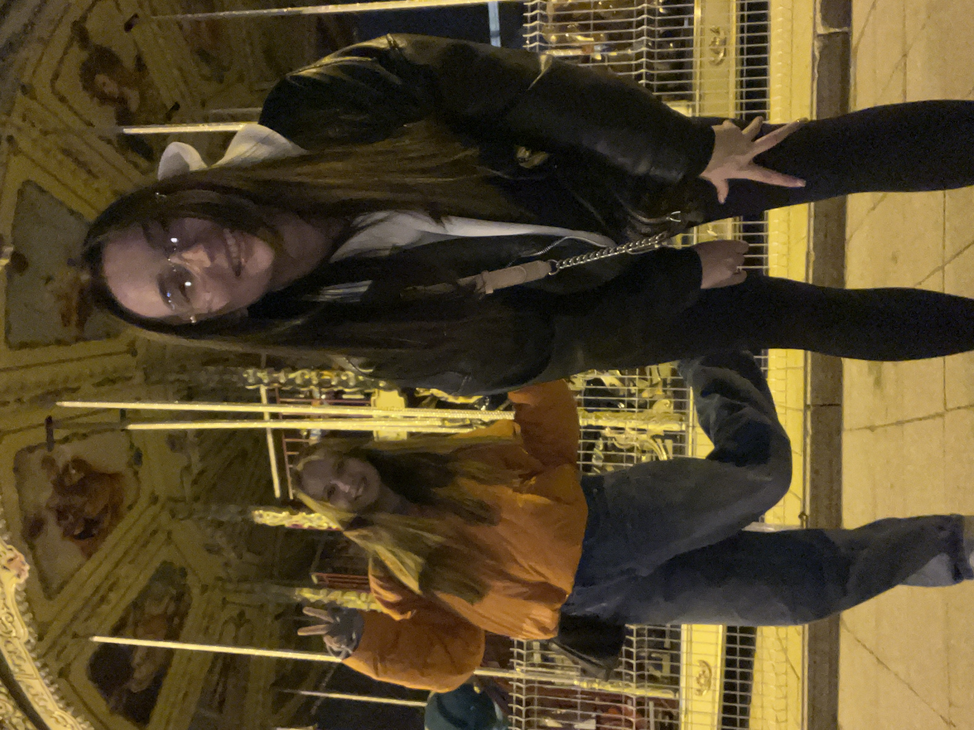 The image depicts two women standing in an outdoor setting at night, likely in Tyumen, Russia, given the presence of a carousel visible in the background. The woman on the left wears a black leather jacket over a white shirt, black leggings, and glasses. She has long, dark hair and a crossbody bag with a chain strap. The woman on the right wears an orange jacket and jeans and is holding up a peace sign. The background features an ornately decorated ceiling that appears to be part of the carousel's structure, indicative of the city's known carousel locations. The lighting is predominantly yellow, suggesting artificial illumination. They are standing next to a metal railing and a light colored sidewalk.