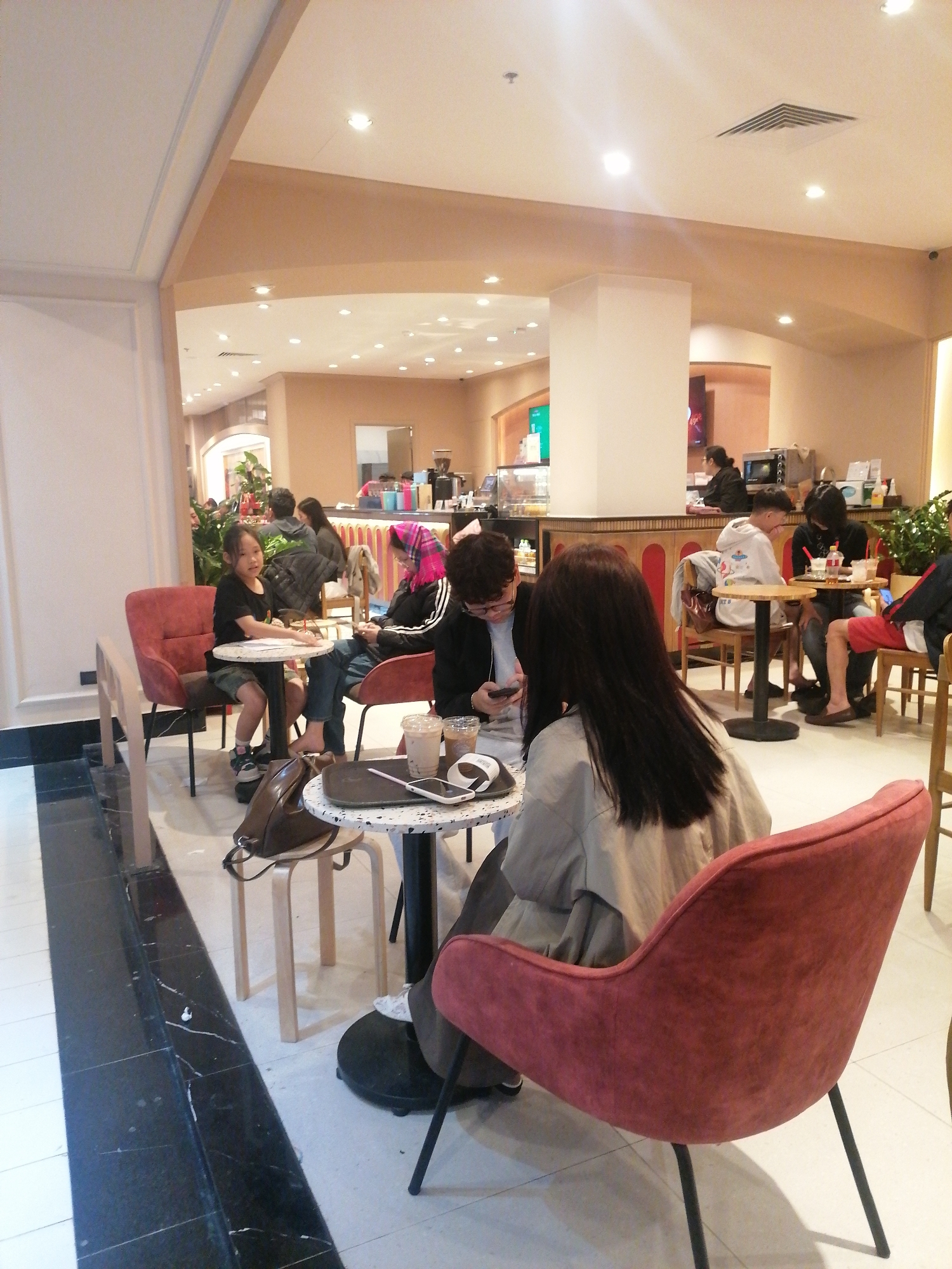 A brightly lit indoor space, possibly a coffee shop, in Sa Pa Ward, Vietnam, contains several people seated at small tables. A young girl sits at a round table with another person, while a person with dark hair in a light-colored jacket sits at a nearby table. On the table are two drinks, a phone, and a white case. More people sit at tables in the background toward a counter area. The space has a modern interior design with light-colored walls and tiled floors.