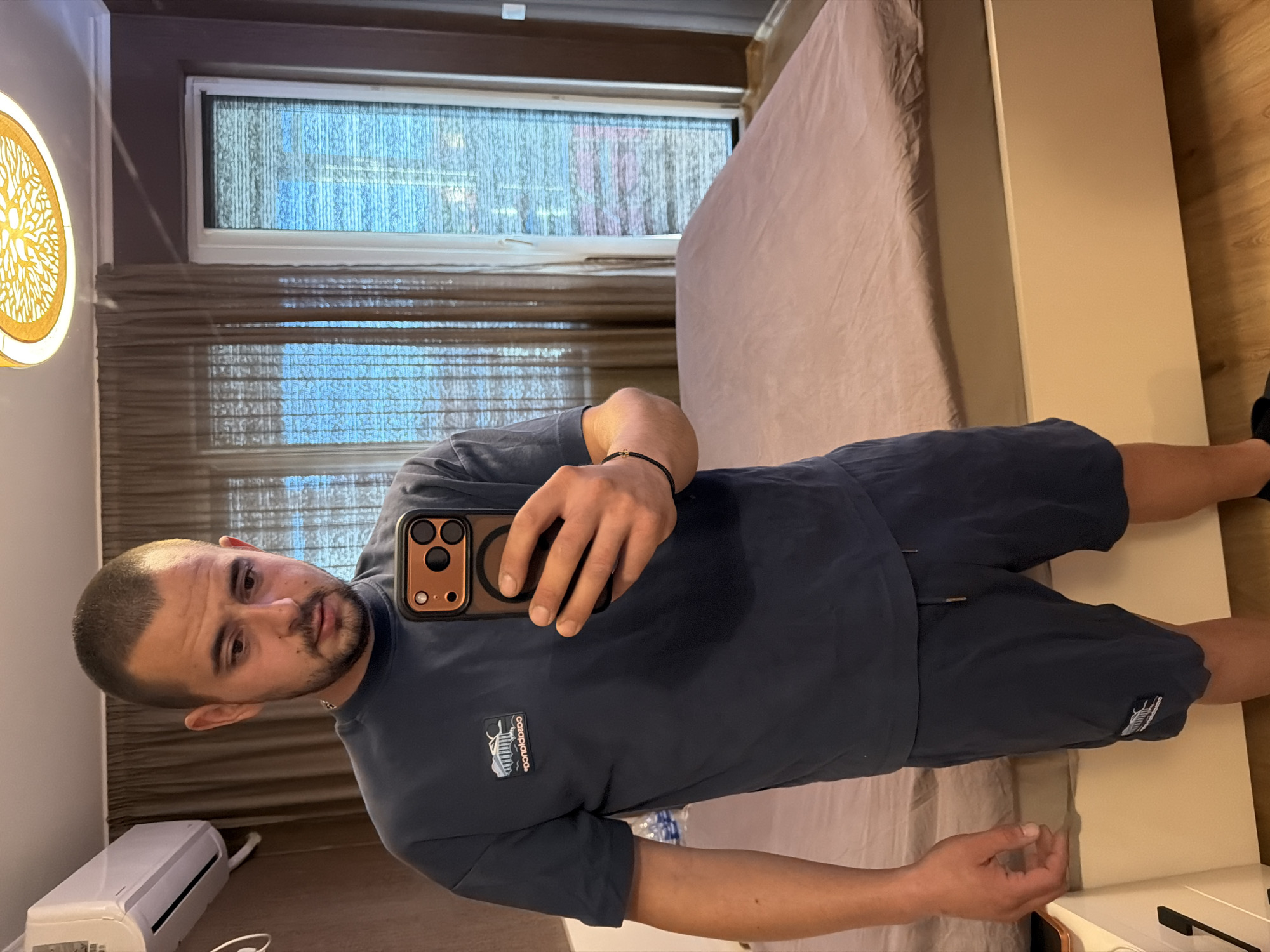 A man with short dark hair and a beard is standing in a bedroom, taking a mirror selfie. He is wearing a dark blue t-shirt with a small logo on the left chest and dark blue shorts. He is holding a smartphone in his right hand, which has a copper-colored case with three black camera lenses. On his left wrist, he wears a thin black bracelet.
The bedroom has light brown walls. To the left, a white air conditioning unit is mounted high on the wall. The room has a large bed covered with a light pinkish-brown duvet. Behind the bed are sheer brown curtains and vertical blinds covering a window. The man's reflection shows that he is standing in front of the bed. A light fixture with a yellow, patterned shade hangs from the ceiling.
The scene is indoors, likely a residential room. No elements in the image directly indicate the city of Burgas, Bulgaria.