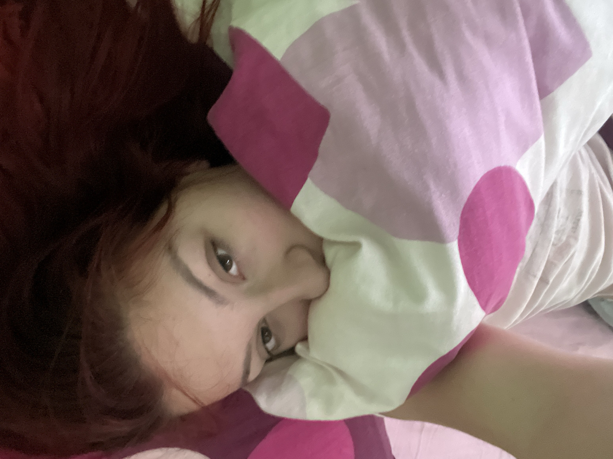 A person with reddish-brown hair is lying in bed, partially obscured by a pillow. The pillow has a pink and white patterned design. The person's face is visible, with brown eyes and some features covered by the pillow. The scene appears to be indoors, possibly a bedroom. There is no indication that the image was taken in Asenovgrad, Bulgaria.