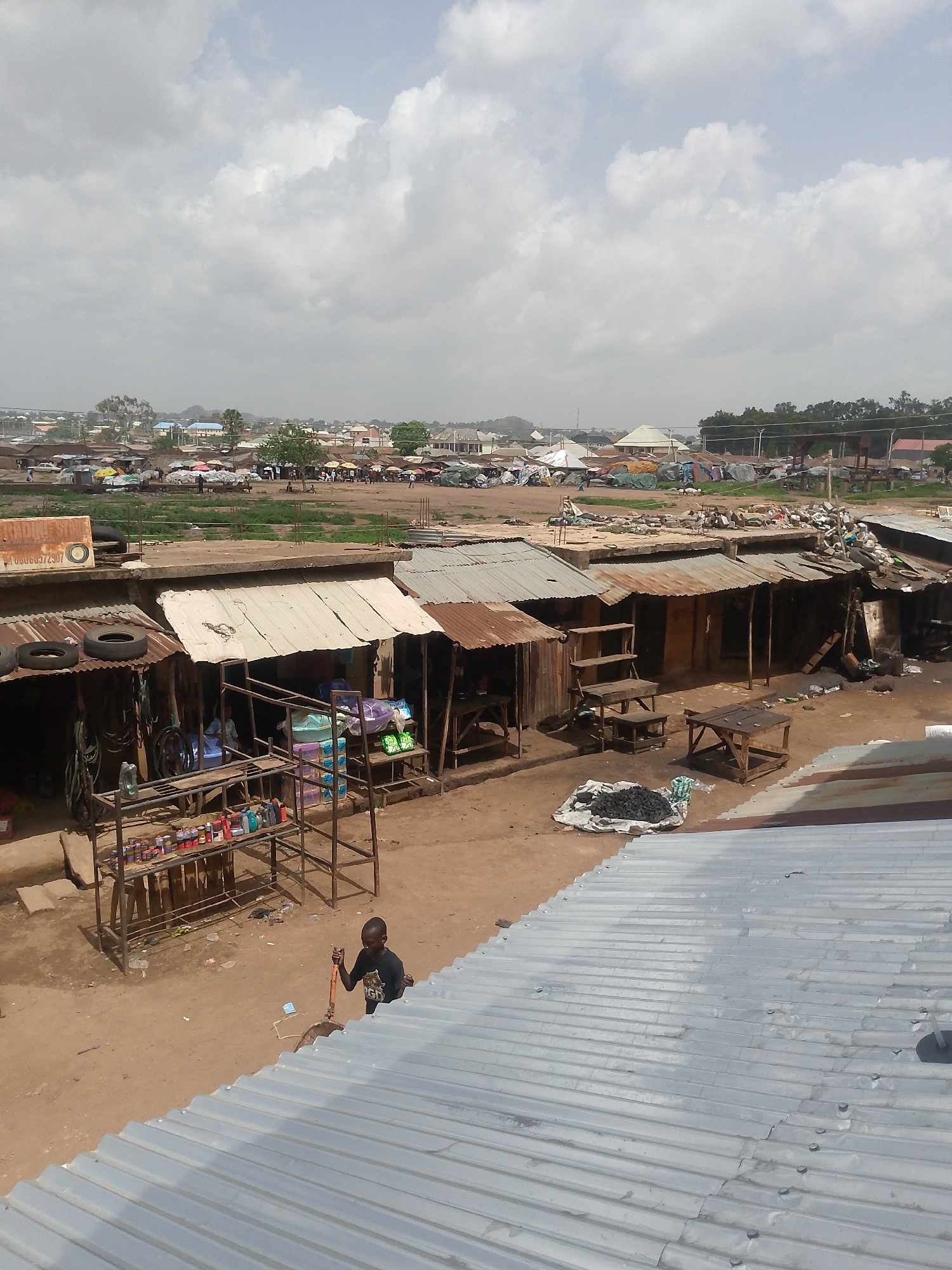 The image depicts a bustling market scene in Bukuru, Nigeria. The foreground shows a corrugated metal roof, possibly the vantage point from where the photo was taken. In the middle ground are a series of rudimentary stalls constructed from wood and corrugated metal sheets, displaying various goods. A young boy stands in the dusty ground between the stalls. The background features an open space with many people and market umbrellas, surrounded by buildings of various sizes and construction. The sky is overcast with scattered clouds. The overall scene suggests a vibrant, though somewhat impoverished, urban environment.