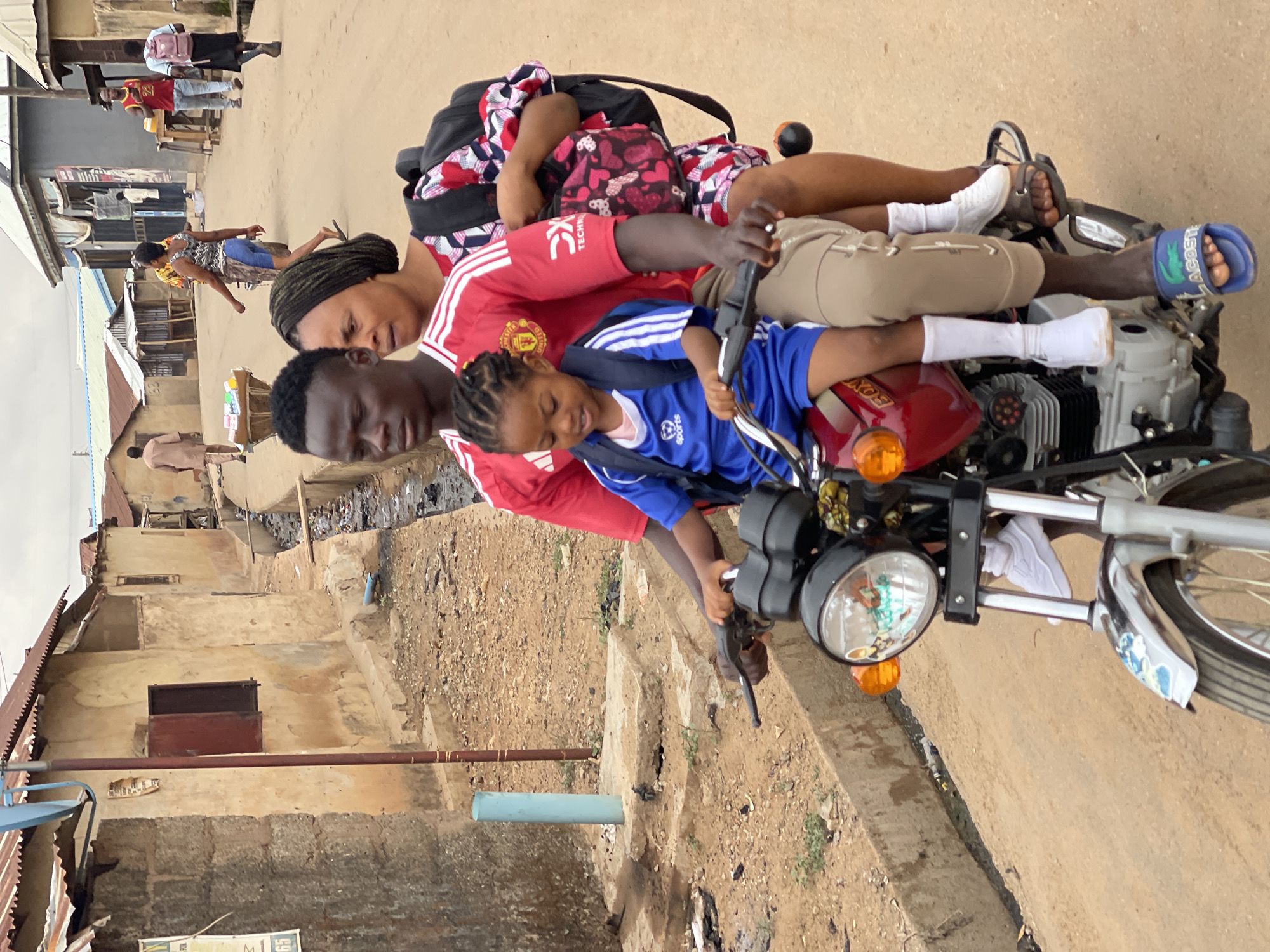 The image depicts a family of three on a motorcycle in Karmajiji, Nigeria. The father, wearing a red Manchester United jersey, is driving. A young girl in a blue sports jersey sits in front of him, appearing to hold the handlebars. The mother, with her hair wrapped, sits behind the father, holding onto him, with a backpack. The motorcycle is a standard model with a red fuel tank and silver engine components. The road appears to be a paved street with a curb and some debris. Buildings of simple construction are in the background. Other people are visible in the distance on the street.
