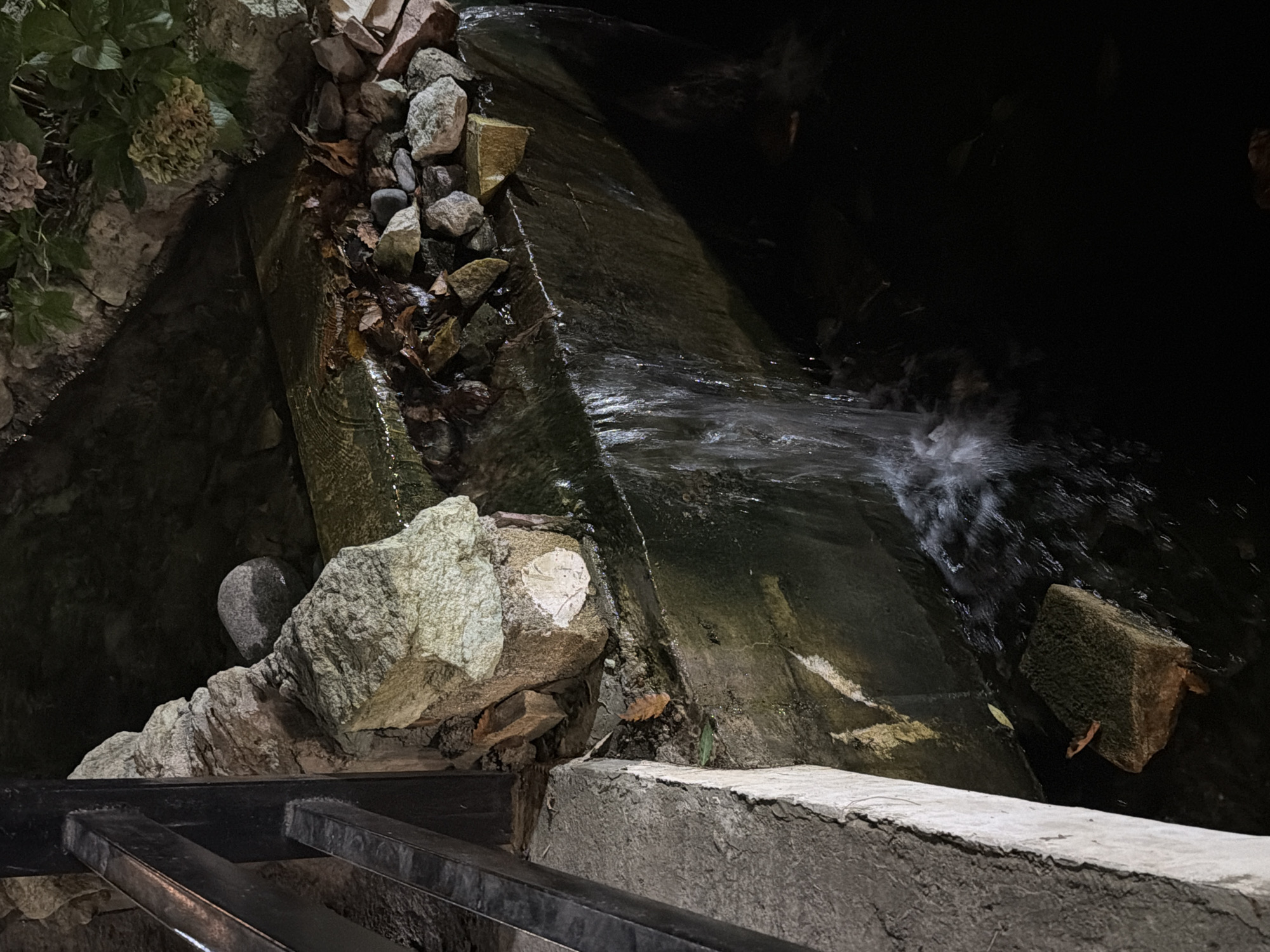 The image shows a man-made waterfall flowing into a dark pool of water. The waterfall is constructed with concrete and rocks, and there are some dry leaves and debris on the rocks above the water. A black metal railing is visible on the left side of the image, adjacent to a concrete ledge. The scene is dimly lit, suggesting it is nighttime. There are no people visible in the image. The location in the city of Qəriblər, Azerbaijan is not discernible from the provided image.