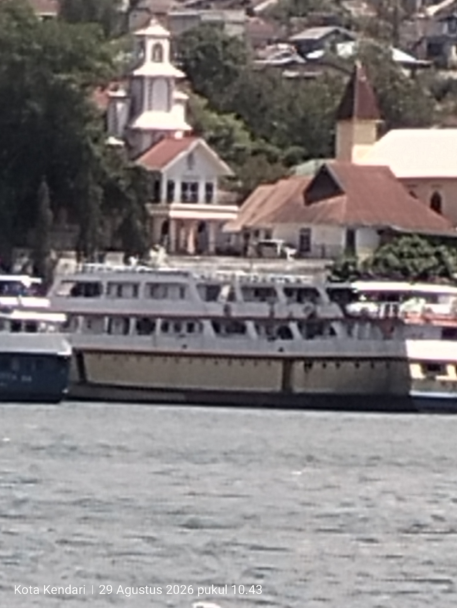 The image shows a body of water in front of a city skyline in Kendari, Indonesia. A large, multi-decked ferry with a yellow hull and white upper decks is docked or moving slowly in the foreground. To the left of the ferry, a smaller dark blue vessel is partially visible. In the background, the cityscape features buildings with red-tiled roofs, some of which appear to be churches or religious buildings, nestled amongst lush green trees and hills. The sky is bright, suggesting daytime. The lower portion of the image contains a text overlay indicating "Kota Kendari | 29 Agustus 2026 pukul 10.43", which translates to "Kendari City | August 29, 2026 at 10:43 AM".