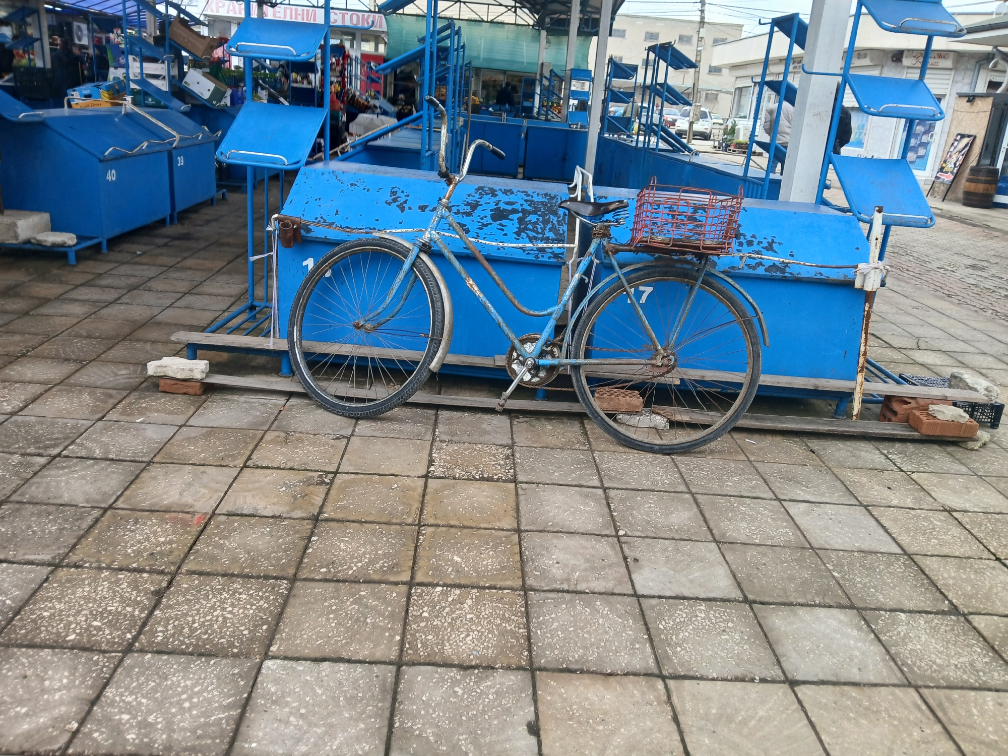 The image depicts a blue bicycle parked in front of blue stalls or containers at what appears to be a market in Kavarna, Bulgaria. The bike has a basket attached to the rear. The stalls are blue and have various numbers and features. The ground is paved with square tiles. In the background, there are more stalls, buildings, and parked vehicles. The lighting suggests an overcast day. There is no visible human interaction.