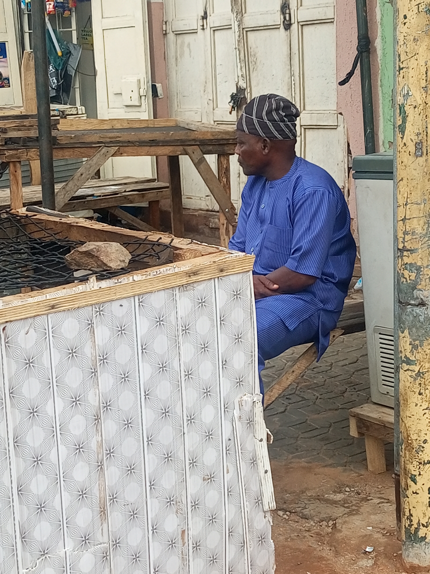 The image depicts an outdoor scene in Unknown, Unknown, featuring one adult male and various makeshift structures and architectural elements.
In the mid-ground, an older adult male with dark skin is seated on a rudimentary wooden bench. He wears a blue, vertically striped traditional outfit, comprising a collared, short-sleeved top with a left breast pocket and matching trousers. A striped cap, black with light grey or white horizontal patterns, covers his head. He is positioned facing left within the frame, with his hands clasped or resting together in his lap.
In the immediate foreground, to the left, is a wooden-framed structure, possibly a stand or grill. Its lower visible section is covered with light-colored panels featuring a repeating geometric pattern resembling starbursts. The top surface of this structure is covered by a black wire mesh, upon which a single irregularly shaped light brown rock is placed.
Behind the man and the foreground structure, a building wall with aged, off-white wooden double doors is visible. A portion of a sign, partially obscured through an opening or window, reads "YELLOW LABEL TEA." A white rectangular electrical box or outlet is mounted on the wall to the right of the doors. A dark green pipe or conduit runs vertically along the wall.
To the right of the seated man, a grey, rectangular appliance, possibly an outdoor air conditioning unit or cooler with a vertical gridded vent at its base, rests on a small wooden block. On the far right, a vertical pole or beam, painted yellow and exhibiting significant paint peeling and weathering, stands against the scene.
The ground surface is a mixture of reddish-brown dirt and dark grey paver stones laid in an interlocking geometric pattern. Various loose wooden planks and construction debris are scattered throughout the scene, contributing to a utilitarian and somewhat disused aesthetic.