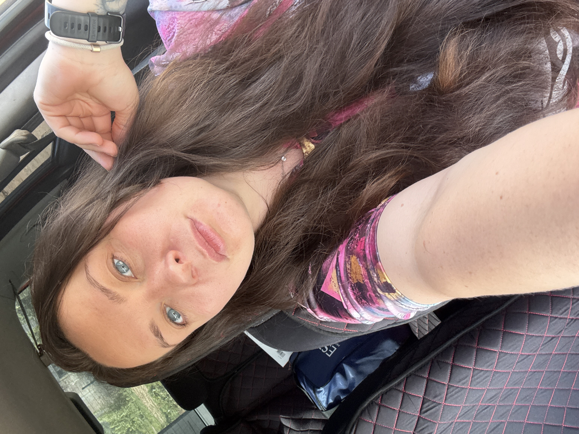 A woman with blue eyes and long brown hair is looking directly at the camera. She is wearing a pink and purple top and a black watch on her left wrist. She is in a car, with the car's interior visible in the background, including the seat and dashboard. A blue bag is partially visible in the lower center of the frame. The background suggests a daytime setting with foliage visible outside the car window. The location in the city of Фоки, Russia cannot be determined from the image alone.