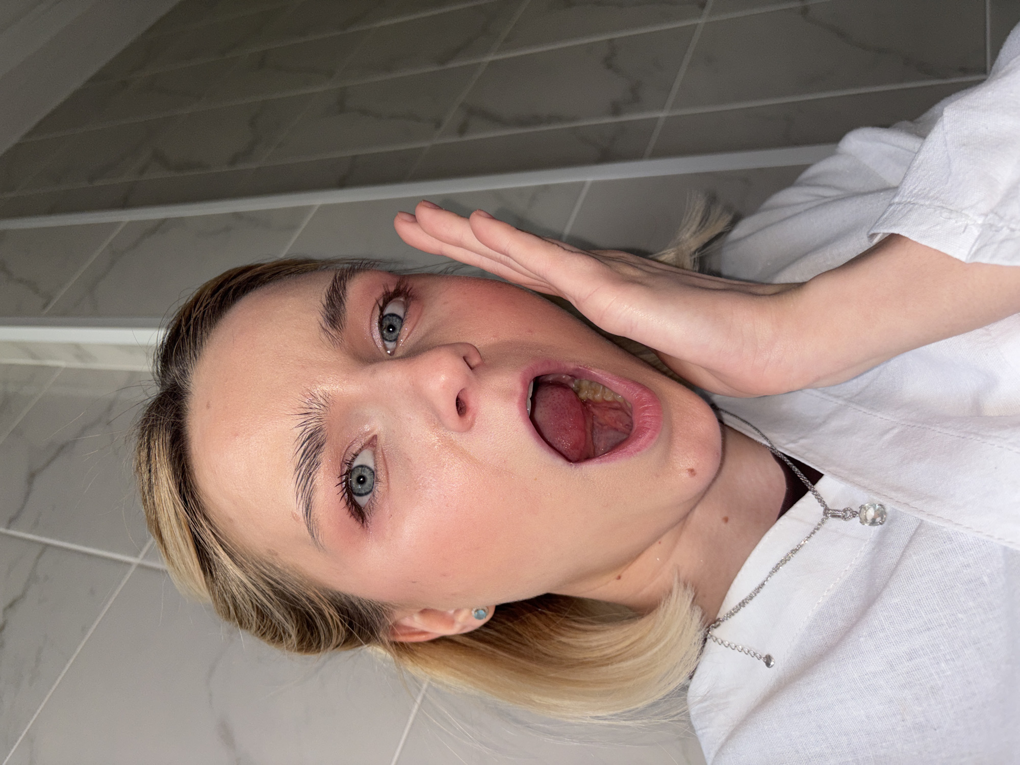 A close-up, rotated portrait features a young person, likely female, with blonde hair and clear blue eyes, depicted indoors. The subject has fair skin and is wearing a white collared shirt, accessorized with a silver necklace featuring a clear, circular pendant and a light blue stud earring in their visible ear.
The person's mouth is wide open, revealing their tongue and the back of their throat, suggesting a loud exclamation, a yawn, or an exaggerated expression of surprise or shock. Their left hand is raised, palm flat against the side of their face, further emphasizing a dramatic or startled reaction. The eyes are directed towards the camera, wide and attentive. The background consists of light grey tiles with subtle darker veining, indicative of a modern indoor setting, possibly a bathroom or a room with a tiled wall. The lighting is bright and even, highlighting the subject's features. There is no visible text in the image. The provided location context is Saint Petersburg, Russia.