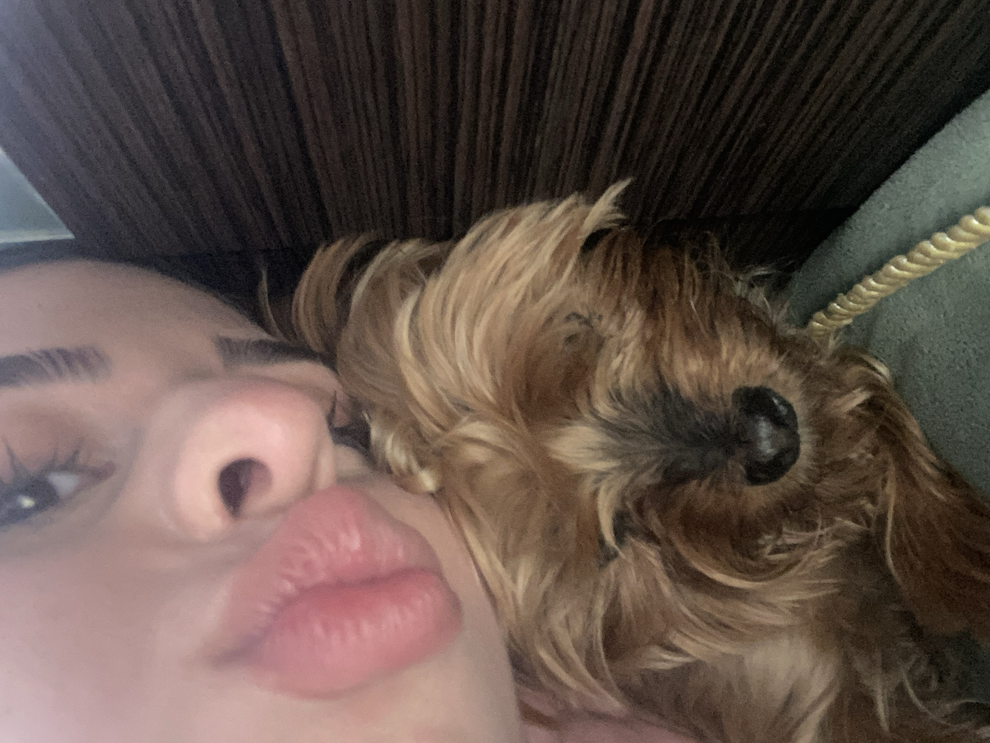 A selfie captures a woman with pursed lips, her face close to the camera, sharing the frame with a brown, furry dog nestled against her. The dog's nose is prominently visible. Above them, a dark, textured surface is seen. To the right of the dog, a green object with a gold rope detail is present. The location is not discernible from the image.