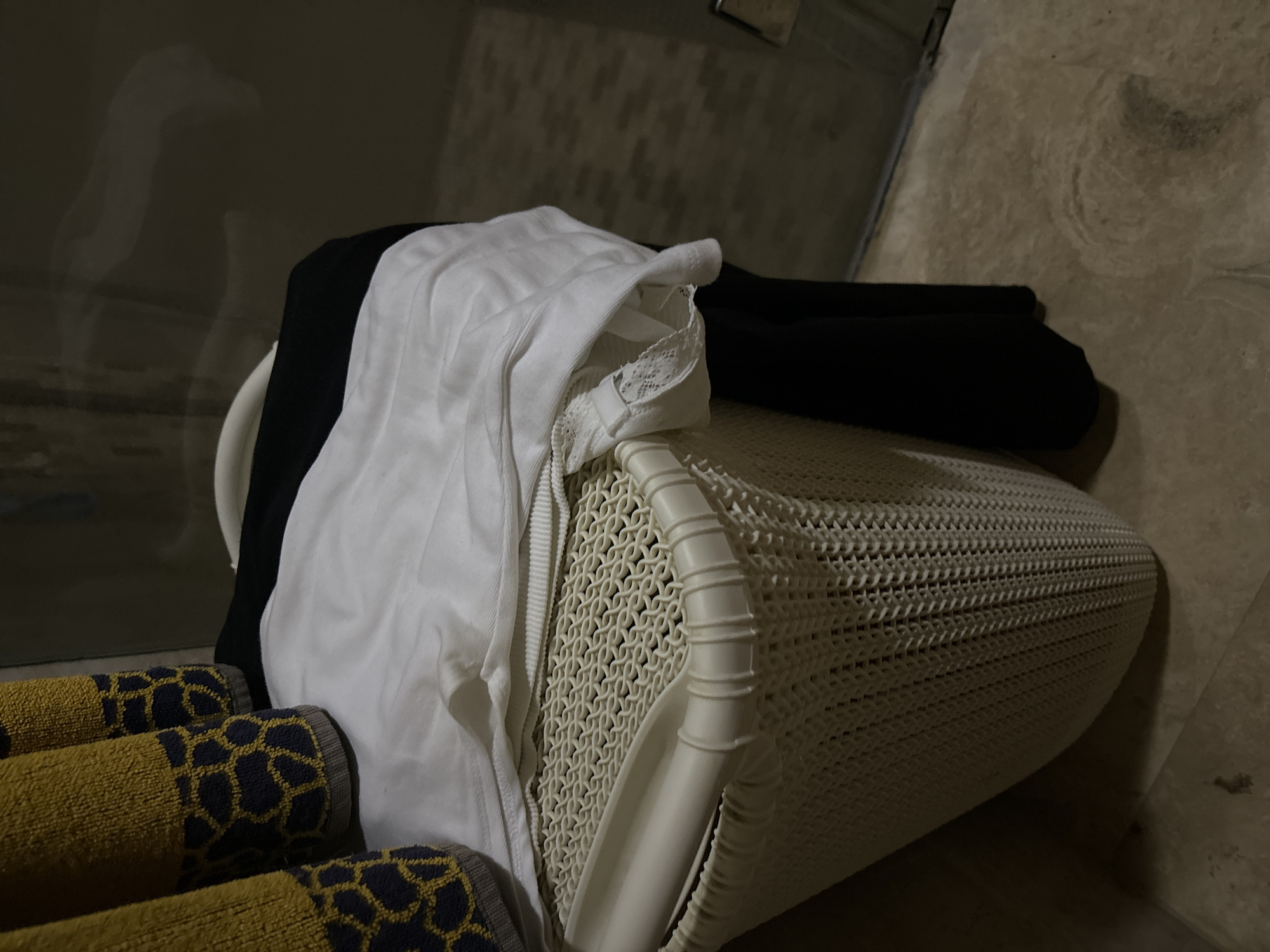 A laundry basket filled with clothes sits on a tiled floor. The basket is white and made of a woven plastic material. On top of the clothes in the basket are two towels with a yellow and navy blue giraffe print. In the background, a glass shower door is visible, and the lighting suggests an indoor environment.