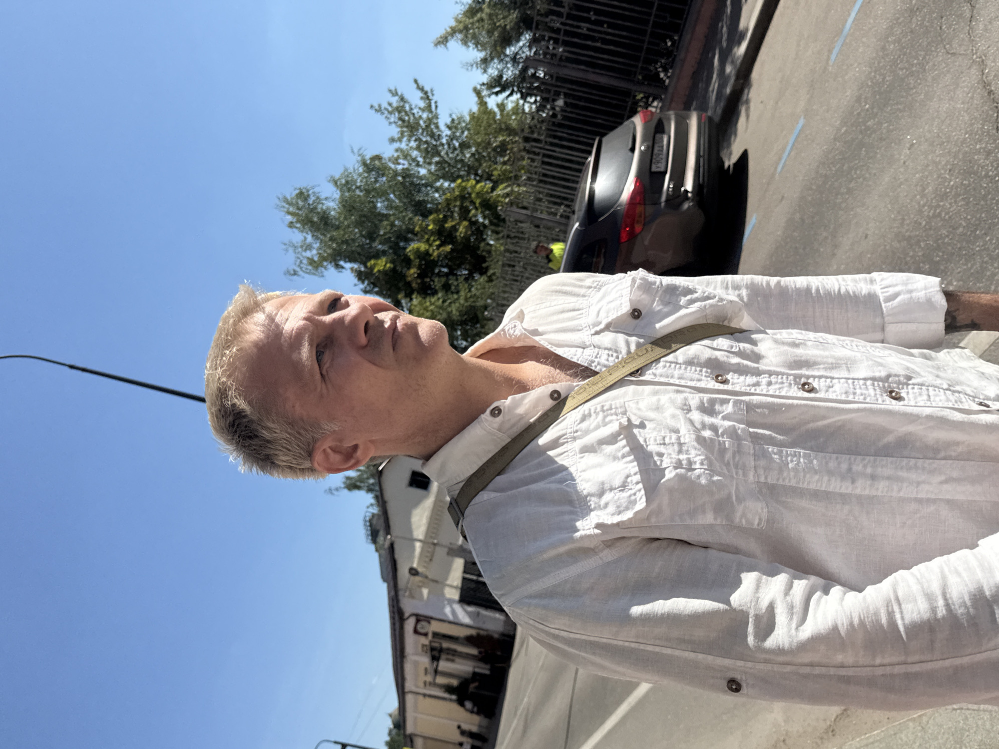 A middle-aged Caucasian man with blond hair and fair skin, wearing a white linen shirt, stands on a street in Moscow, Russia. He is looking to the right of the frame, with a neutral expression. A light green strap is slung across his chest. In the background, a dark brown SUV is parked on the side of the road, and a black metal fence runs along the property line. A few buildings and trees are visible further in the distance. The sky is clear and blue. The image appears to be taken on a sunny day.