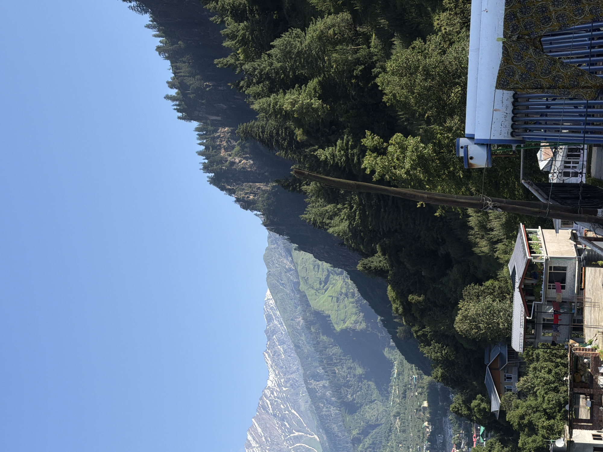 The image depicts a mountainous landscape with a clear blue sky. In the foreground, there are buildings with flat roofs and some laundry hanging. A solar water heater is visible. The midground features a dense forest of pine trees covering a steep, rocky slope. In the background, a series of majestic mountains rise, with snow-capped peaks in the distance. The scene suggests a location in the Indian Himalayas, likely in or near Vashisht, India, known for its natural beauty and thermal springs.