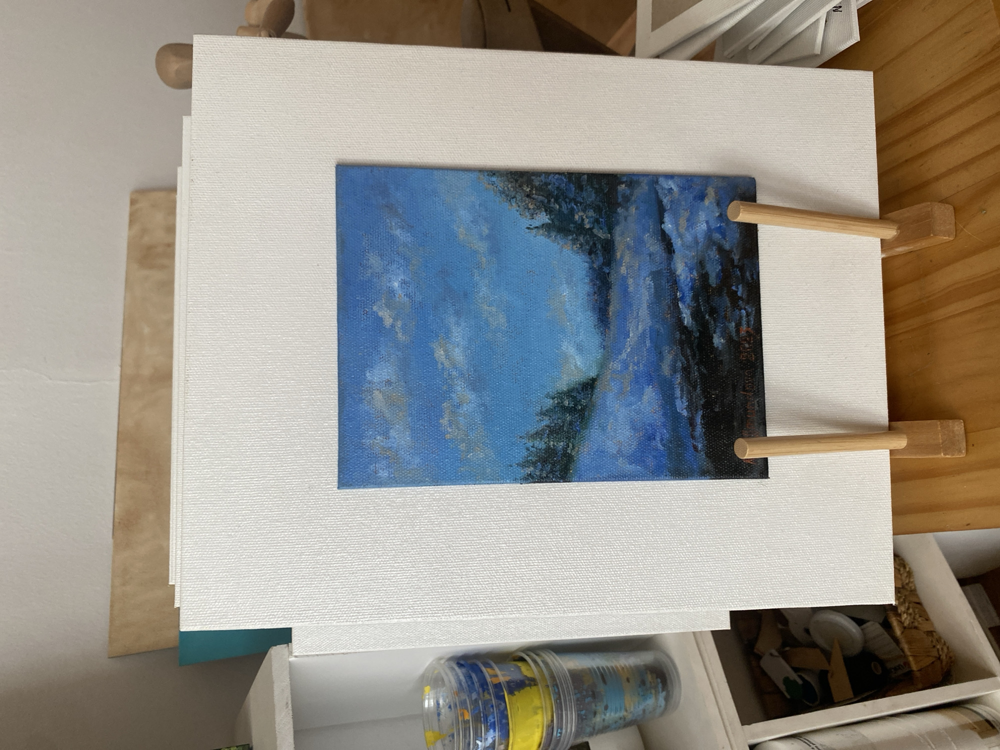 The image shows a small, vibrant oil painting of a snowy landscape with pine trees and a cloudy blue sky, displayed on a wooden easel. The painting is positioned in front of a larger, blank white canvas, with other canvases stacked behind it. To the left, a white shelf unit holds several clear plastic cups filled with colorful paint supplies. The scene appears to be an artist's studio or workspace. The location is not specified.