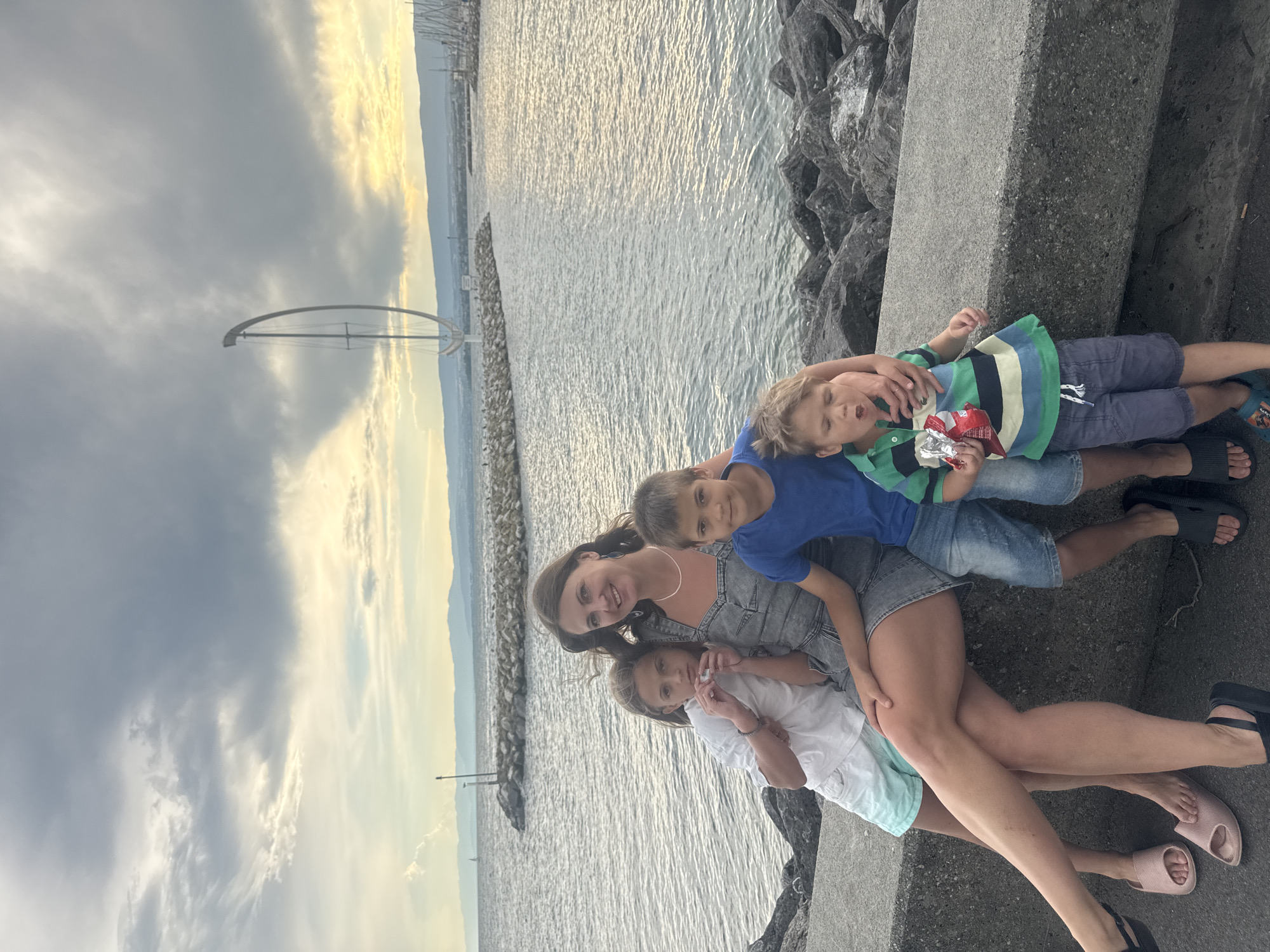 A family of four, a mother and three children, are posing for a photograph while sitting on a concrete barrier by a body of water. The mother is in the center, wearing a denim dress, and is surrounded by her children. To her left is a girl with blonde hair, wearing a white t-shirt and turquoise shorts, and to her right are two boys, one in a blue t-shirt and denim shorts, and the younger one in a striped green and blue shirt and gray shorts. The younger boy is holding a bag of chips. The background features a large body of water, a breakwater made of rocks, and in the distance, a large modern sculpture resembling a half-circle on a stand. The sky is cloudy with patches of sunlight breaking through, suggesting either dawn or dusk. The scene appears to be the Ouchy promenade in Lausanne, Switzerland, identifiable by the Jet d'Eau sculpture which is a prominent landmark.
