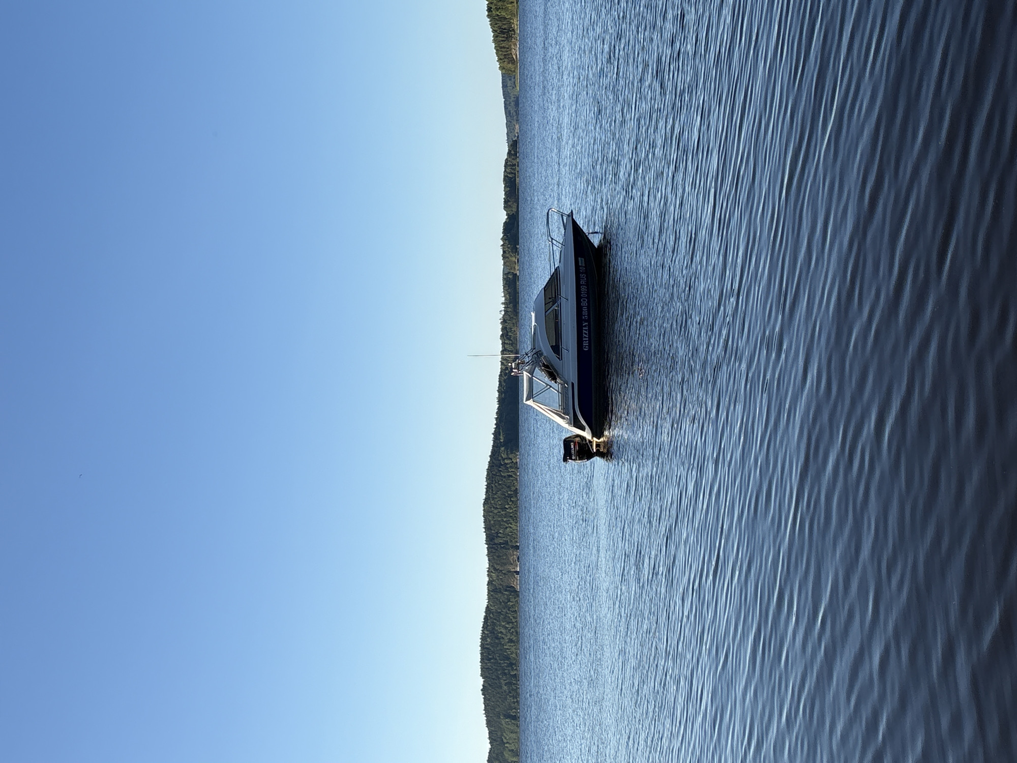 A white and blue motorboat with a Suzuki outboard motor is anchored in calm, blue water. The boat has a covered cabin and a railing around the deck. The boat is inscribed with "GRIZZLY 580 BO 0199 RUS 10". In the background, a dense, tree-covered shoreline stretches across the horizon under a clear blue sky. The scene is characteristic of the natural environment near Rautalakhti, Russia, likely a serene lake or bay. No people are visible in the image.
