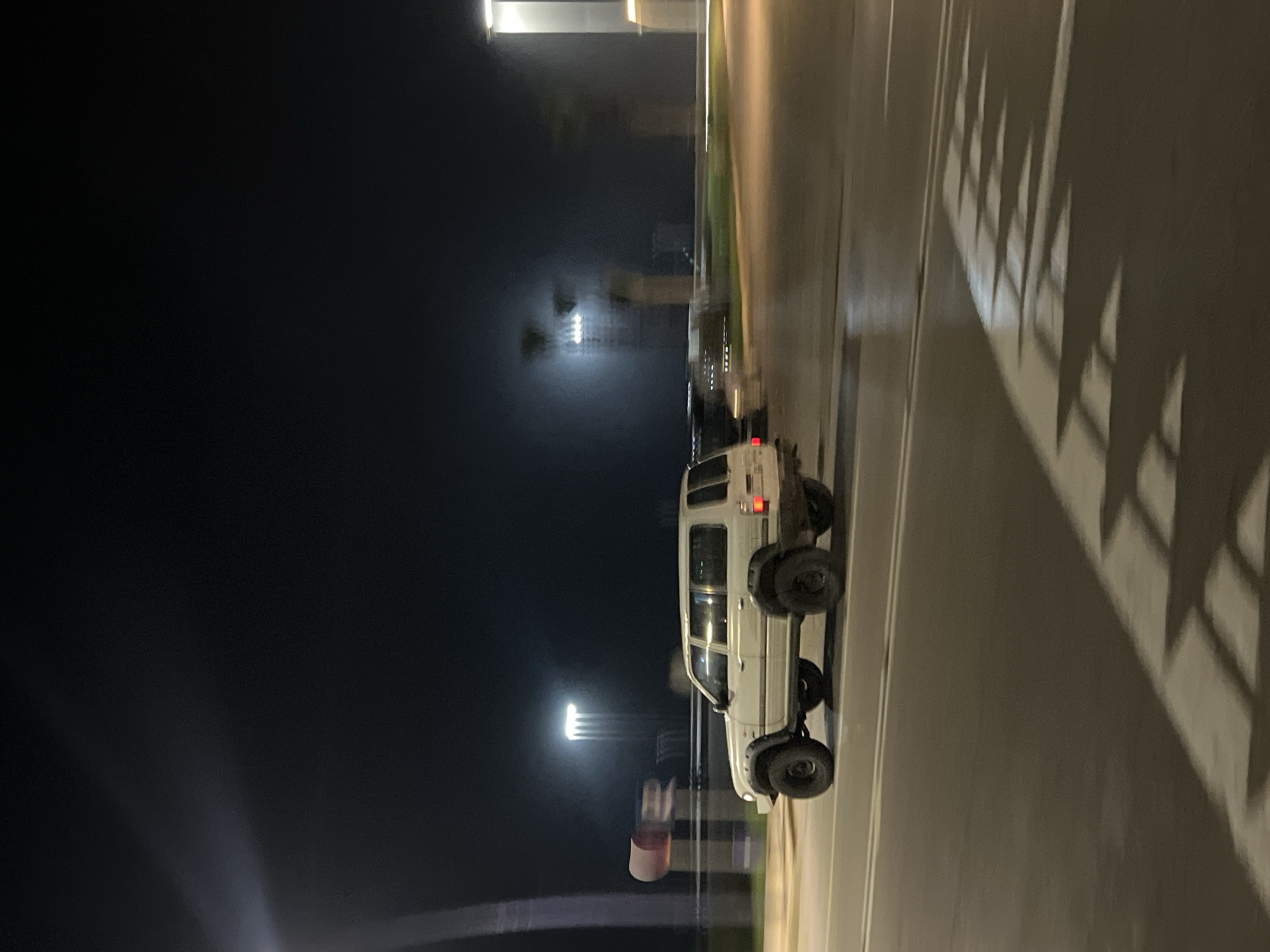 The media file captures a night scene on a paved road. A white sport utility vehicle (SUV), likely an older model with robust, possibly off-road tires, is prominently featured on the right side of the road. Its rear lights are illuminated.
The time of day is clearly night, characterized by the dark sky and the presence of artificial illumination. Streetlights cast bright glows in the distance and create long, sharply defined shadows on the road surface. These shadows, resembling a railing or fence, extend from the right foreground towards the vehicle. The background is dark and somewhat blurry, showing distant lights that suggest buildings or other structures in an urban or semi-urban environment. There are no people visible, and no specific activity is taking place other than the car being present on the road, possibly parked or moving slowly. No visible text is discernible in the image. The scene is set in M'Diq, Morocco.