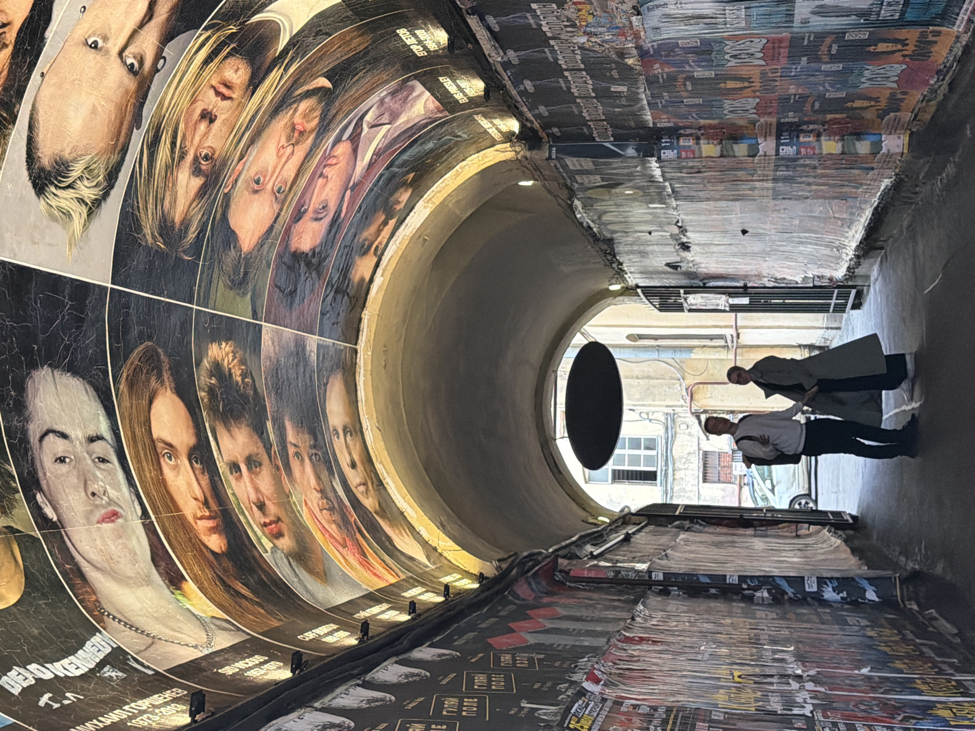 The image depicts an archway with the ceiling adorned with a collage of portraits in a curved, mosaic-like fashion. The walls on either side of the archway are covered in numerous posters, some of which are torn and peeling, creating a layered effect. Two individuals are standing in the middle of the archway, facing away from the camera and towards the end of the passage. The person on the left is wearing a white long-sleeved shirt, dark pants, and a backpack. The person on the right is wearing a long, light-colored coat and dark pants. The scene appears to be an outdoor urban setting, possibly a pedestrian alley or a passageway between buildings. The lighting suggests it is daytime.