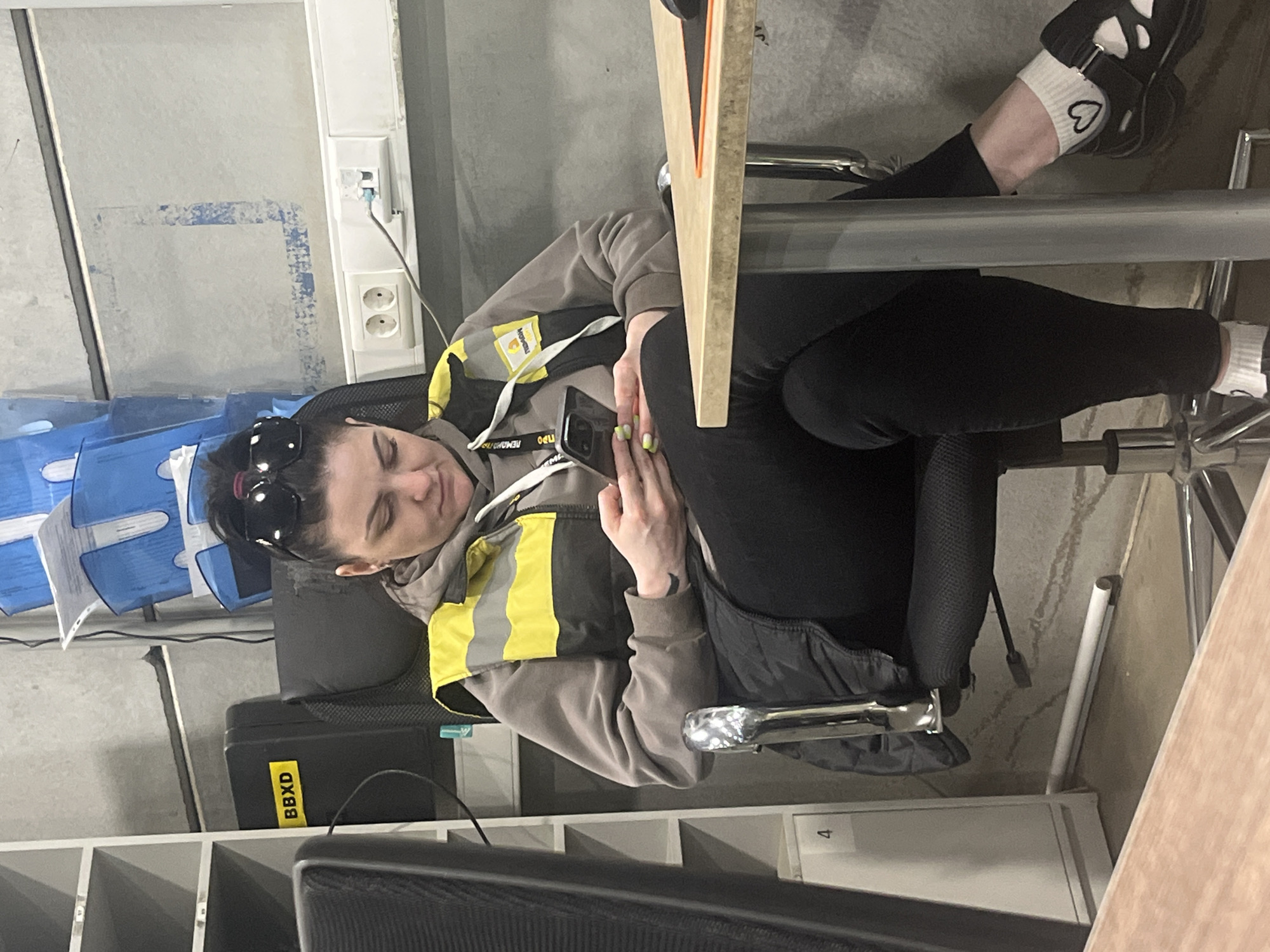 A woman reclines in an office chair, using a desk as a makeshift headrest. She is wearing a grey hooded sweatshirt with a yellow and grey high-visibility vest over it, black leggings, white socks with a black heart design, and black sandals. Sunglasses are perched on her head. She is holding a smartphone in her hands.
To the left are stacked blue plastic trays and a black equipment case labeled "BBXD." In the background is a window and a wall with electrical outlets. There are shelves to the lower left. The setting appears to be an office environment. There is no indication of location in Lupanovo, Russia.